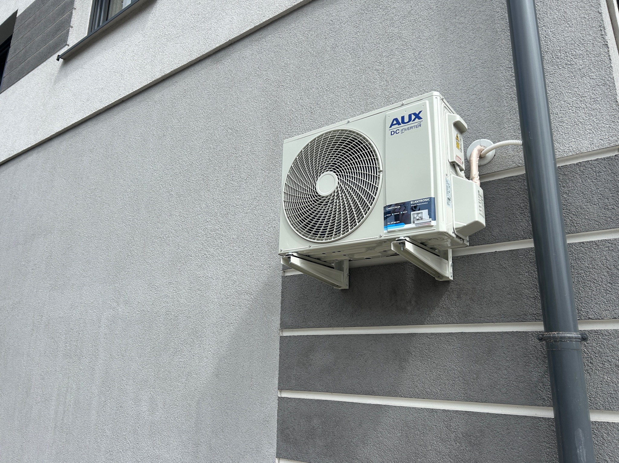 Biała jednostka zewnętrzna klimatyzacji AUX DC Inverter zamontowana na szarej, teksturowanej ścianie budynku z widocznymi elementami instalacji.