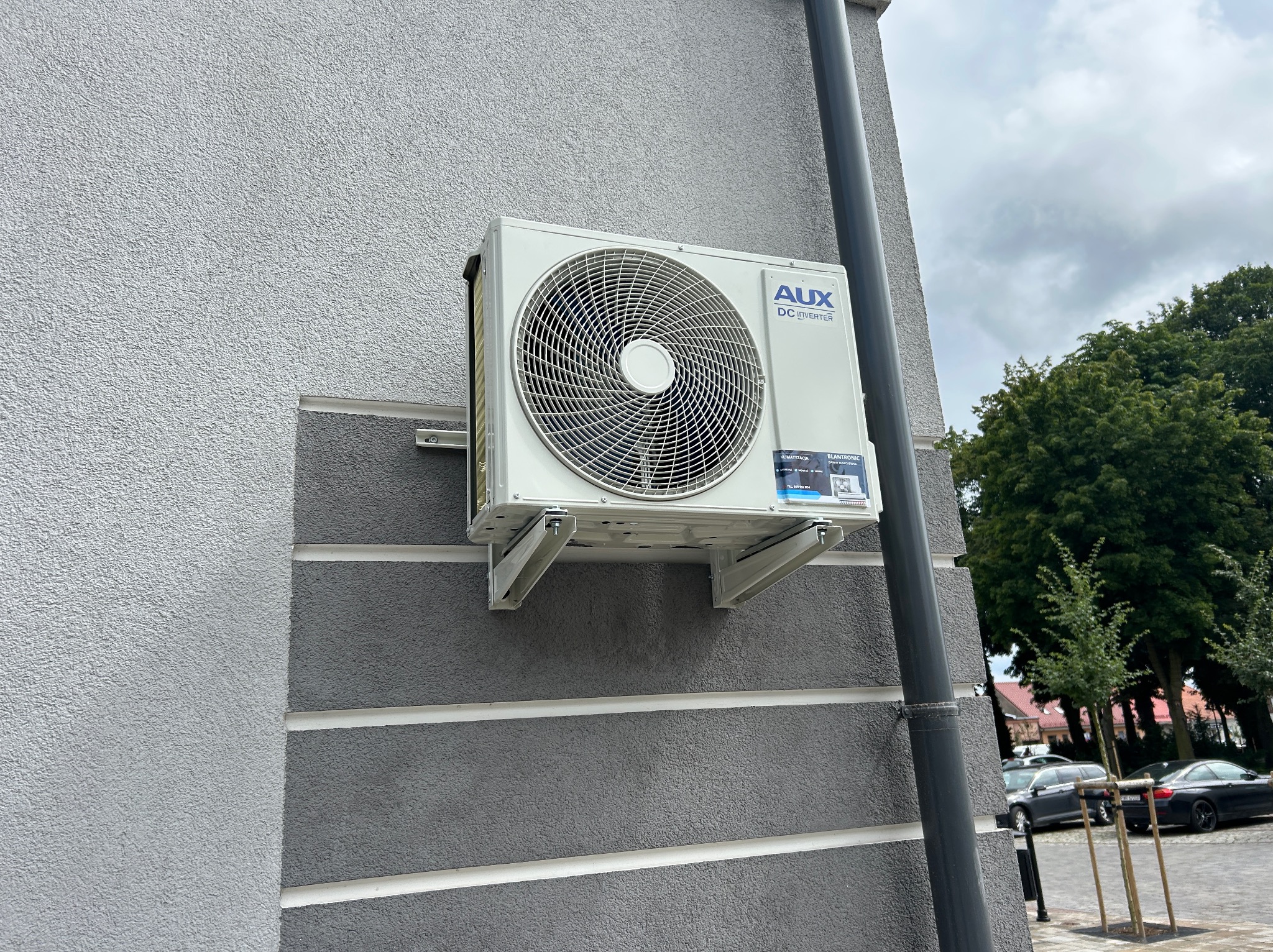 Biała jednostka zewnętrzna klimatyzacji AUX DC Inverter zamontowana na szarej elewacji budynku z widocznymi elementami montażowymi i fragmentem otoczenia.