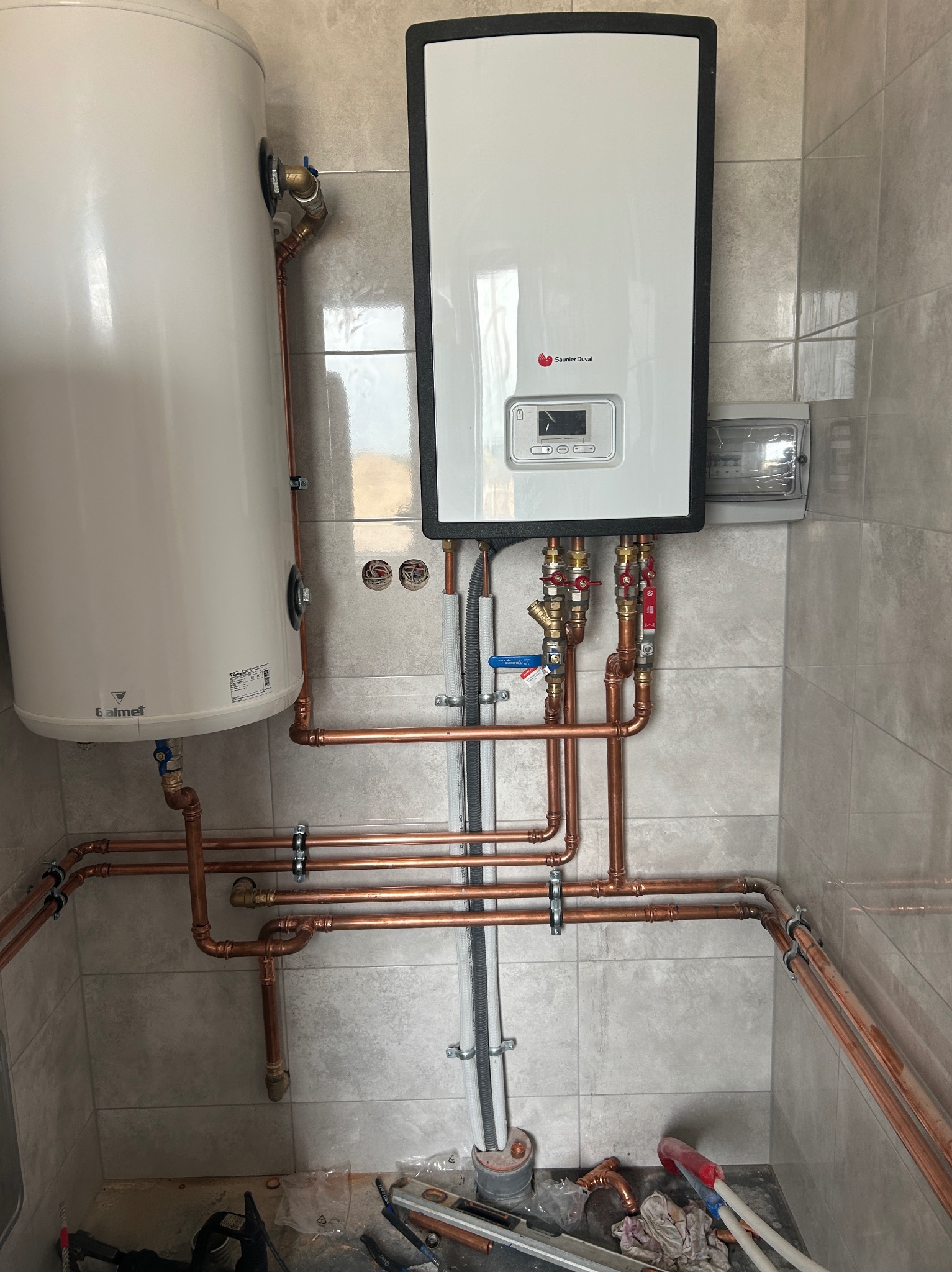 Biały bojler i piec gazowy Saunier Duval z miedzianą instalacją na tle szarych płytek. Widoczne narzędzia instalacyjne na podłodze.