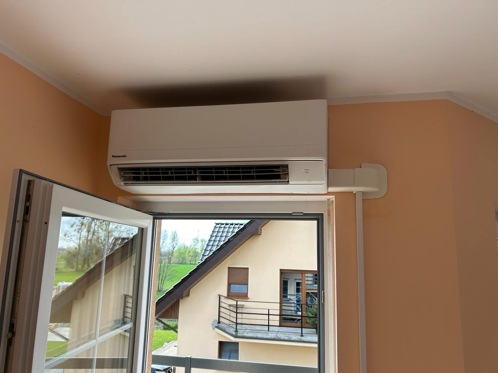 Biała klimatyzacja Panasonic zamontowana nad oknem z widokiem na dom z balkonem. Instalacja z maskownicą na ścianie w kolorze brzoskwiniowym.