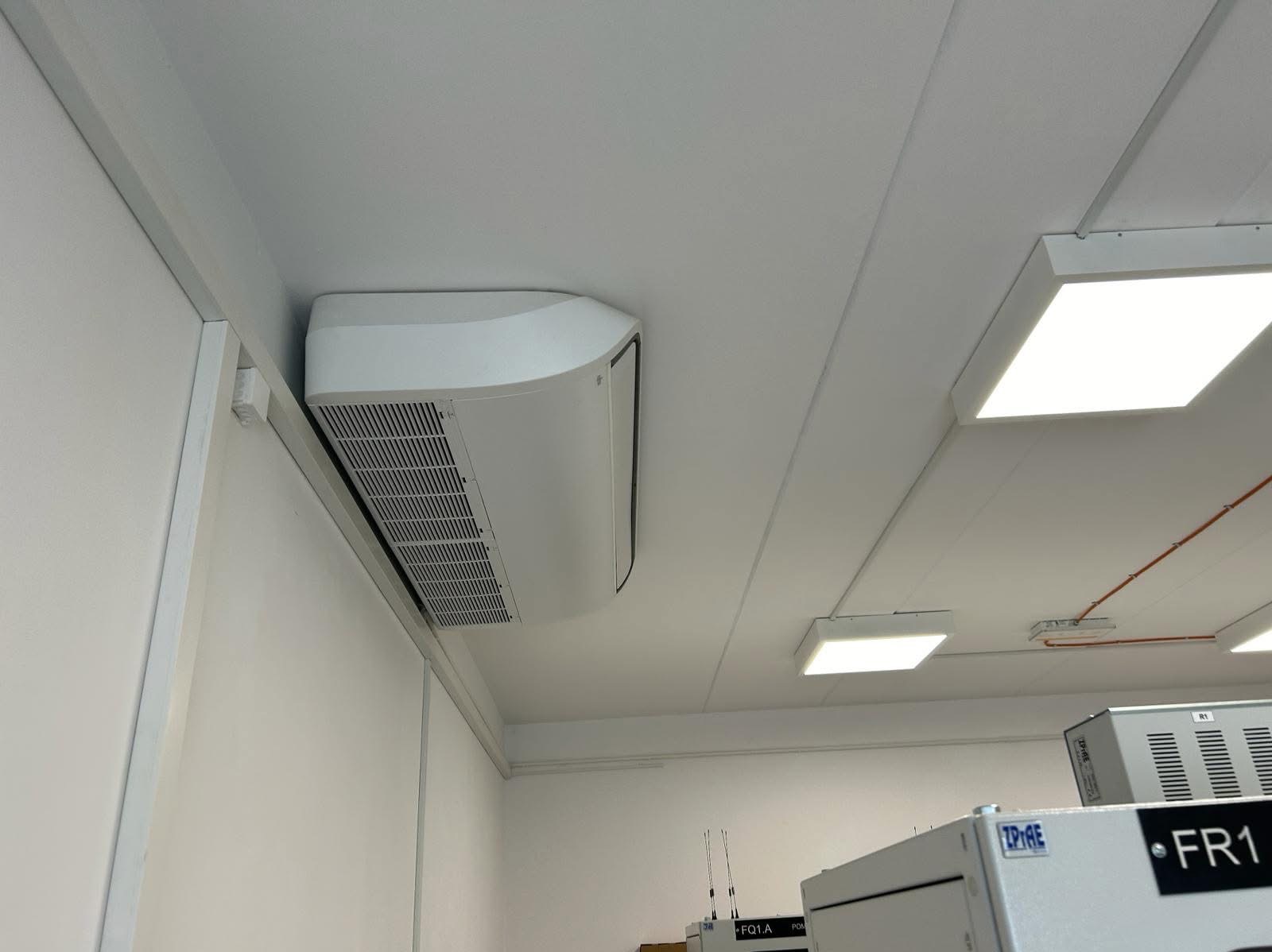 Biała, sufitowa klimatyzacja przemysłowa zamontowana w pomieszczeniu biurowym z oświetleniem LED. Widoczne elementy instalacji i inne urządzenia biurowe.