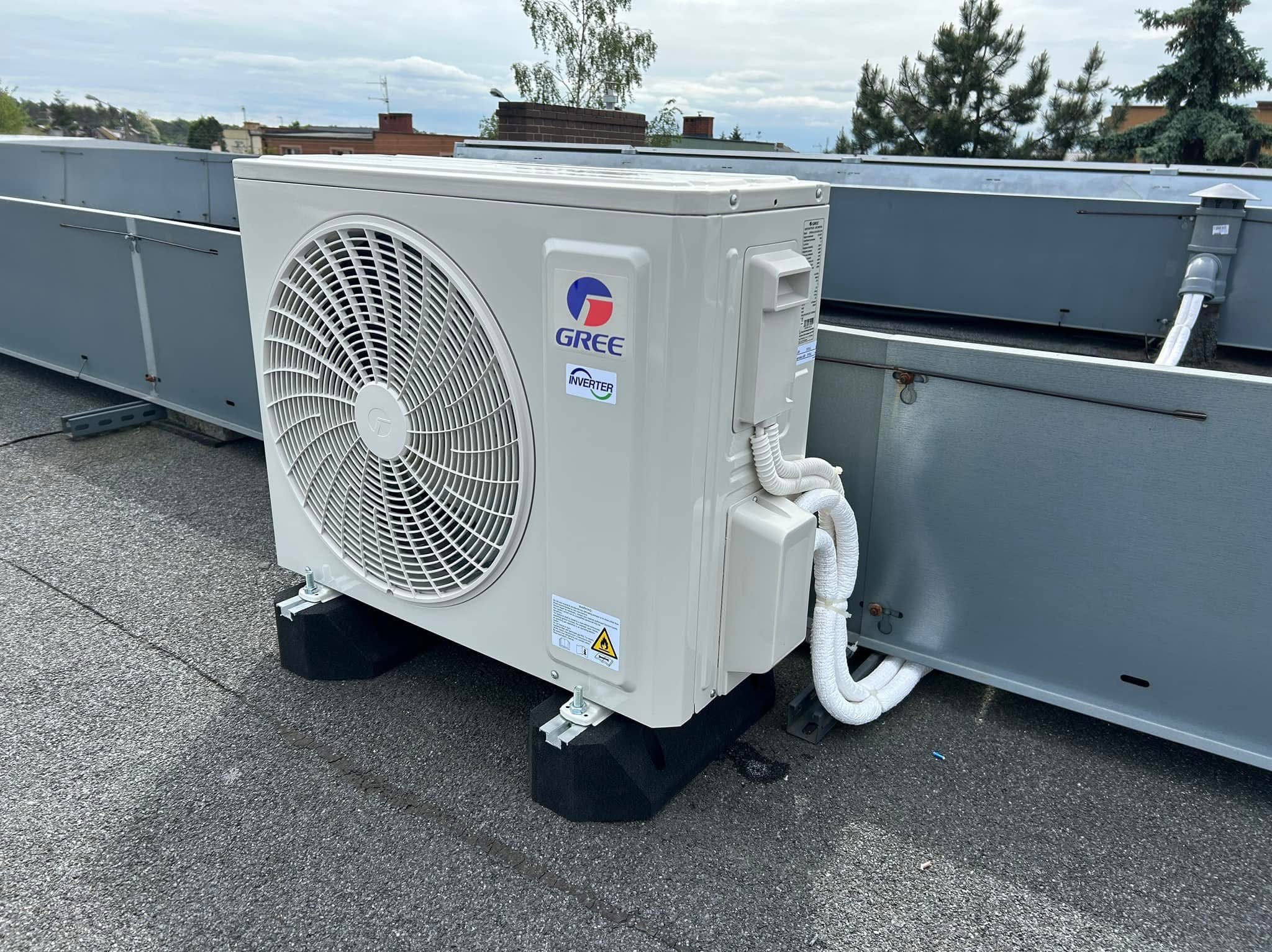 Biała jednostka zewnętrzna klimatyzacji Gree Inverter na dachu, zamontowana na czarnych podkładkach antywibracyjnych. Widoczne przewody i elementy instalacji.