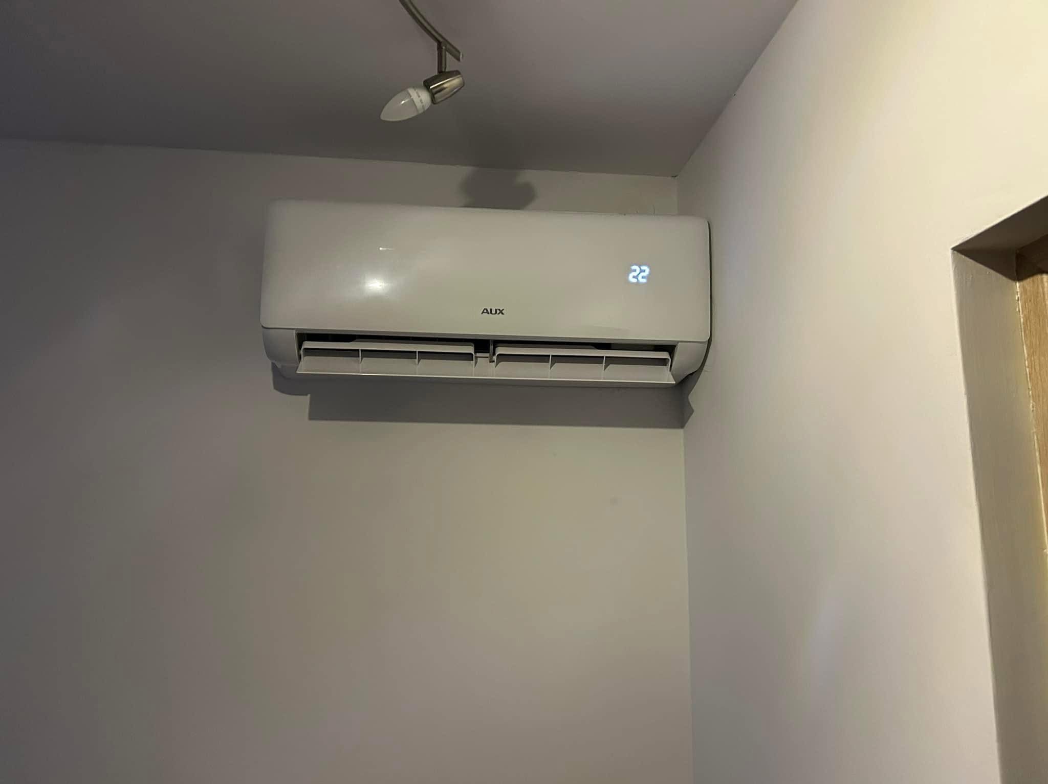 Biały klimatyzator AUX zamontowany na ścianie w pomieszczeniu, wyświetlający temperaturę 22 stopnie Celsjusza. Widoczna lampa sufitowa i fragment futryny drzwi.