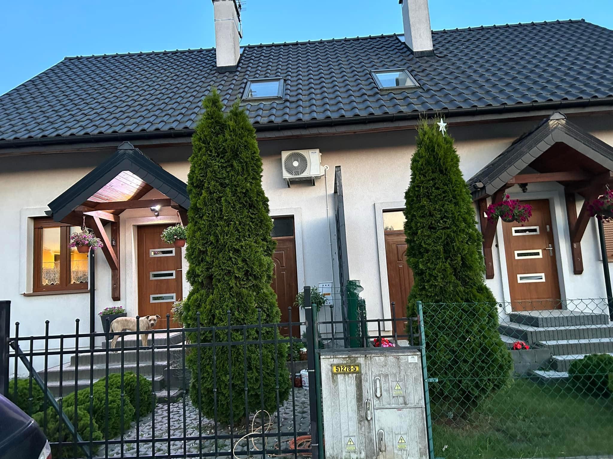 Biały dom z klimatyzatorem na ścianie, widoczny zza ogrodzenia. Dwa wysokie tuje po bokach, dach z oknami połaciowymi. Pies na schodach wejściowych.