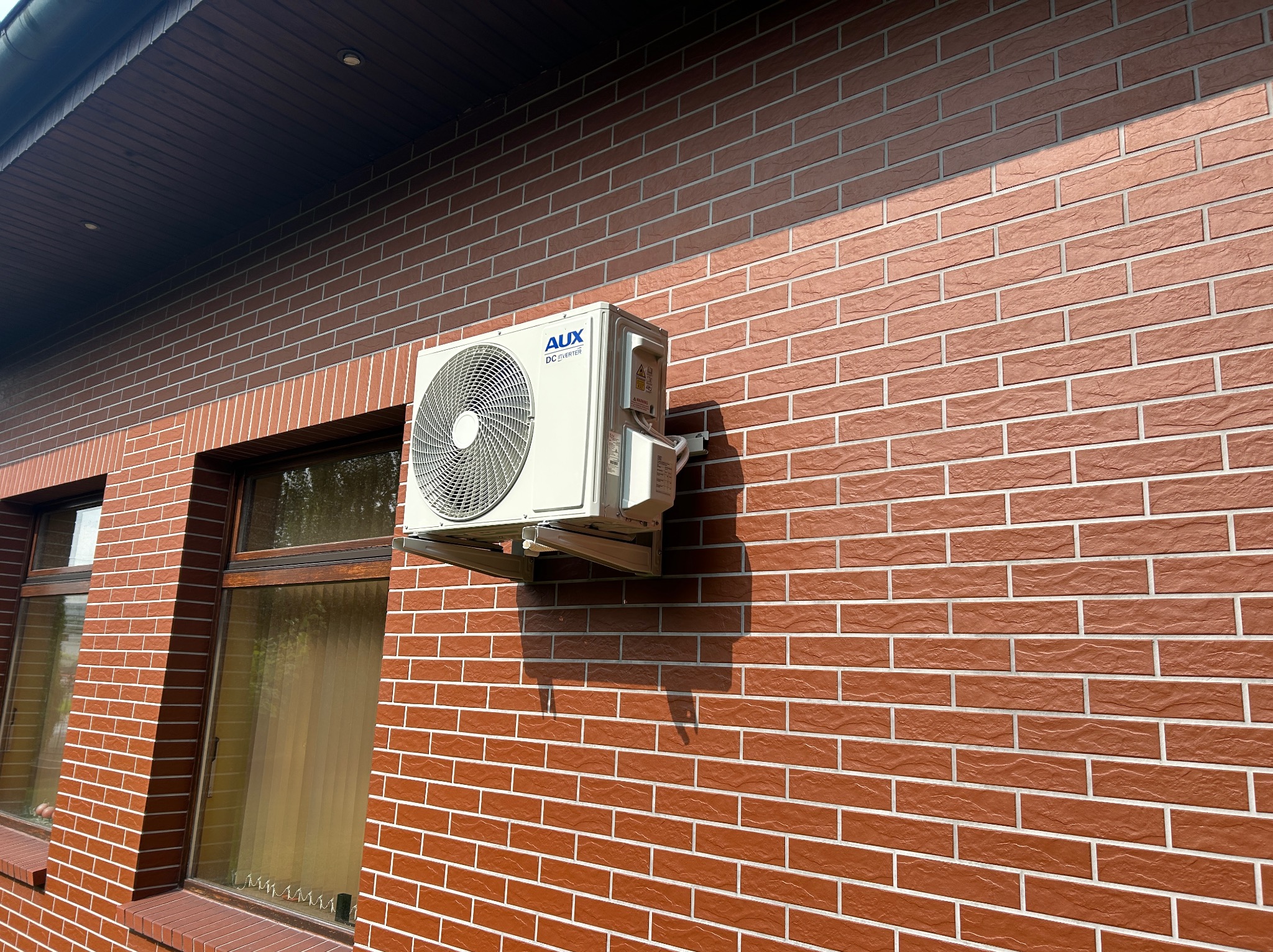 Klimatyzator AUX DC Inverter zamontowany na elewacji budynku z cegły klinkierowej w Zagórowie. Widok z boku, cień urządzenia na ścianie.
