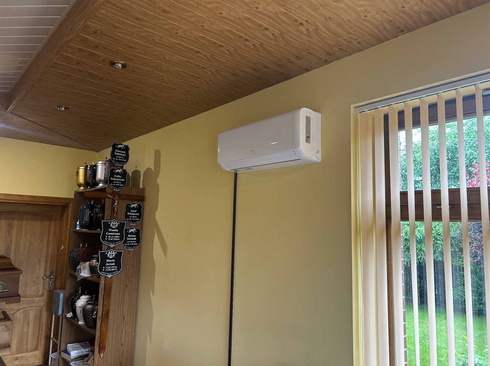 Biały klimatyzator ścienny AUX zamontowany w pokoju z drewnianym sufitem i żaluzjami pionowymi. Widoczny kabel zasilający. Nietypowe otoczenie.