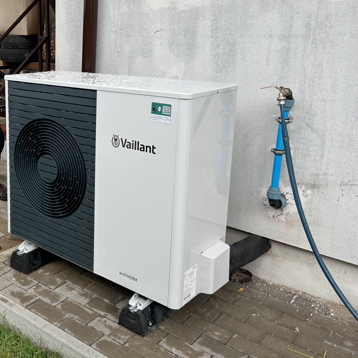 Biała pompa ciepła Vaillant aroTHERM na zewnątrz budynku, z widocznymi przyłączami hydraulicznymi i elementami montażowymi. Świeżo po instalacji, na mokrej kostce brukowej.