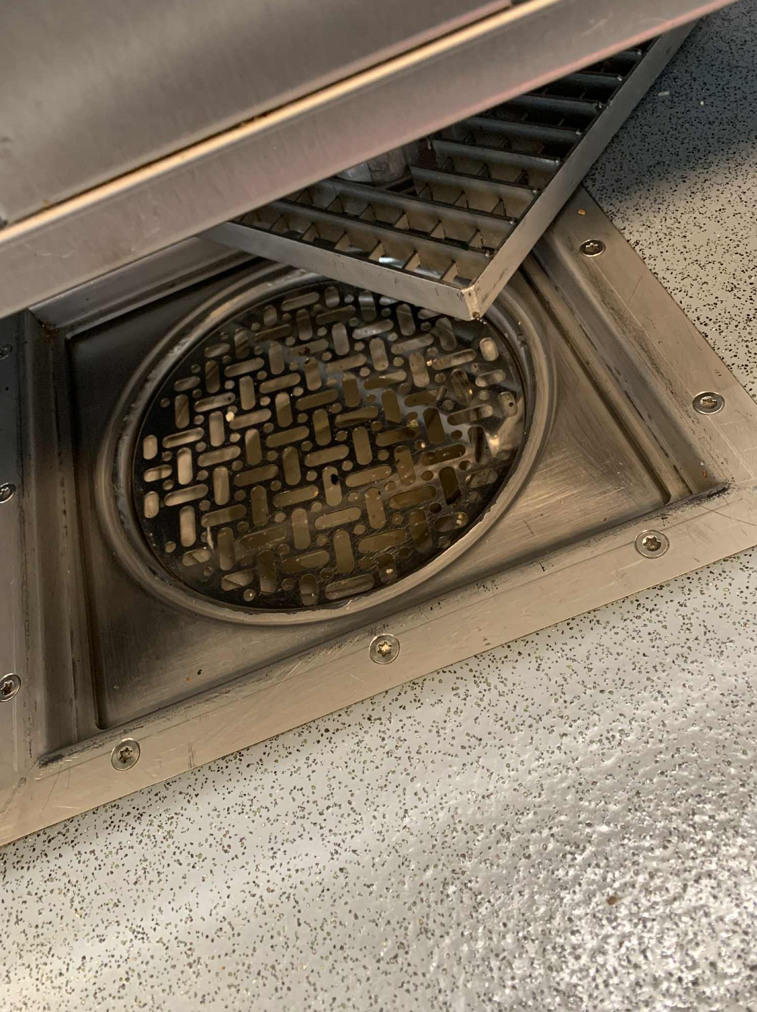 Otwarty, metalowy odpływ podłogowy z widoczną kratką i osadami, umieszczony w jasnej, nakrapianej posadzce. Częściowo uniesiona kratka odpływu.