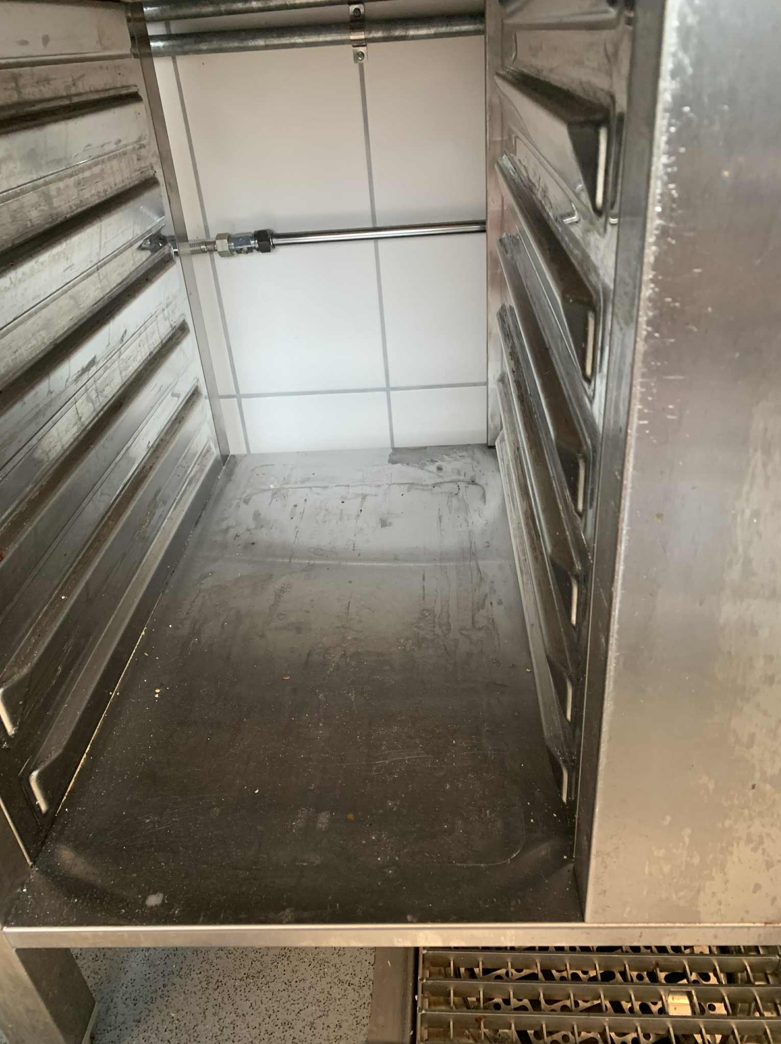 Wnętrze brudnej, metalowej komory piekarniczej z widocznymi prowadnicami na blachy i zabrudzeniami na dnie. Białe płytki na ścianie w tle.