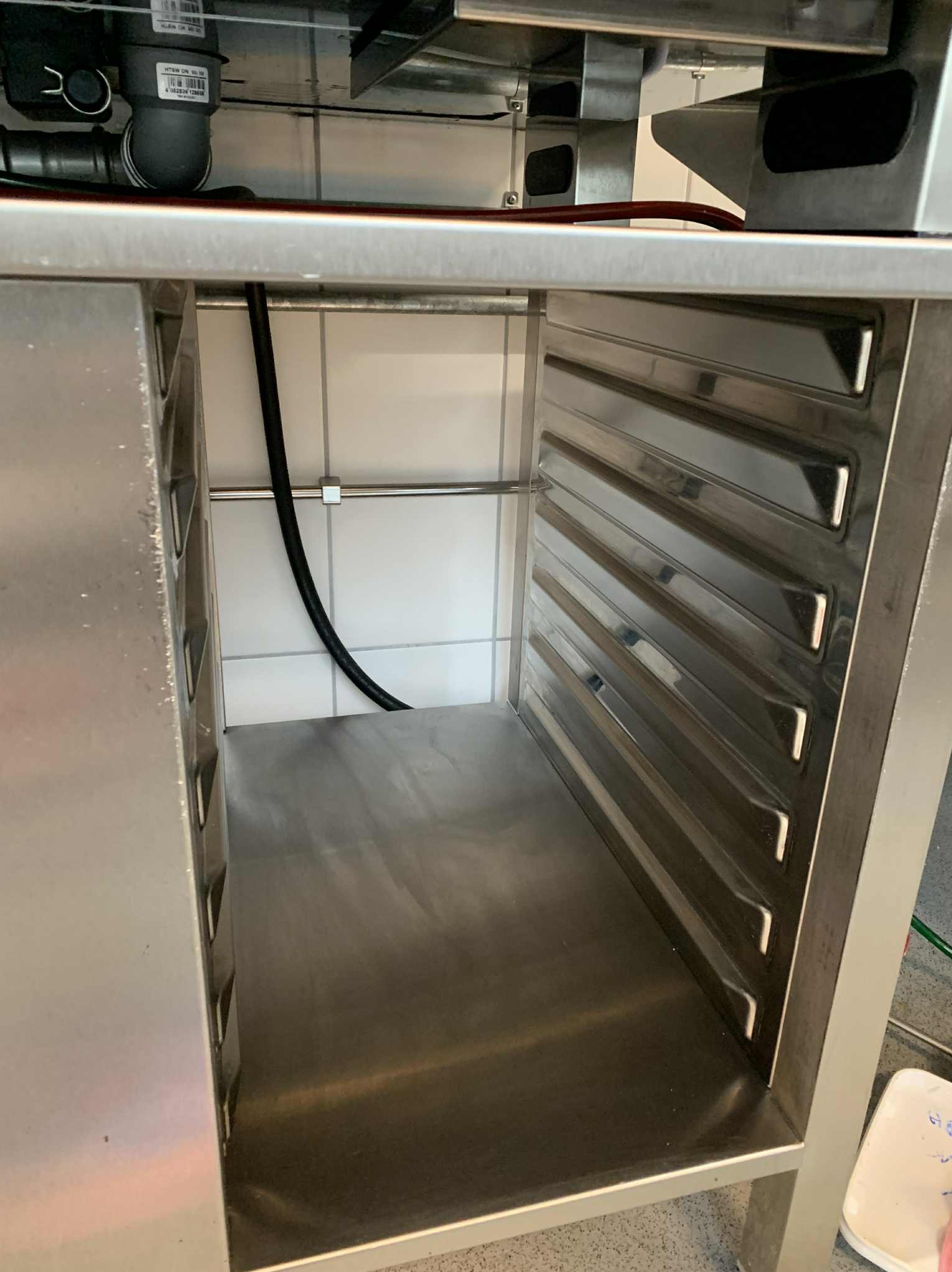 Wnętrze profesjonalnej, metalowej szafy gastronomicznej z widocznymi prowadnicami na blachy, umieszczonej na tle białych płytek. Widoczny przewód zasilający.