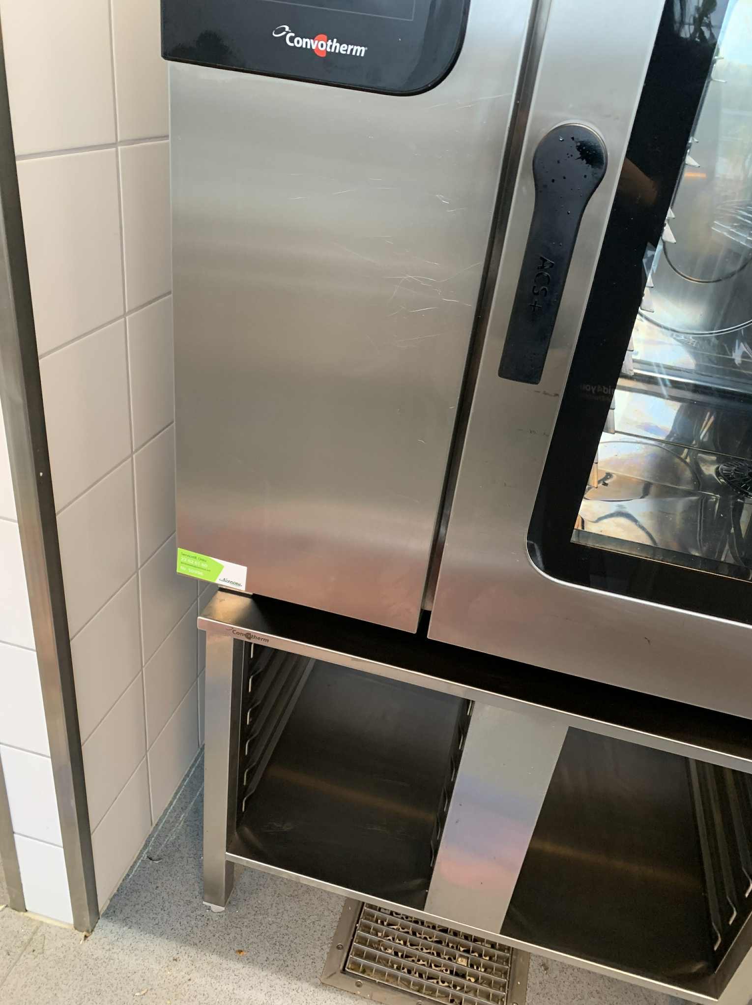 Profesjonalny piec konwekcyjno-parowy ConvoTherm ze stali nierdzewnej, stojący na metalowym regale w pomieszczeniu wyłożonym białymi płytkami. Widoczny odpływ podłogowy.