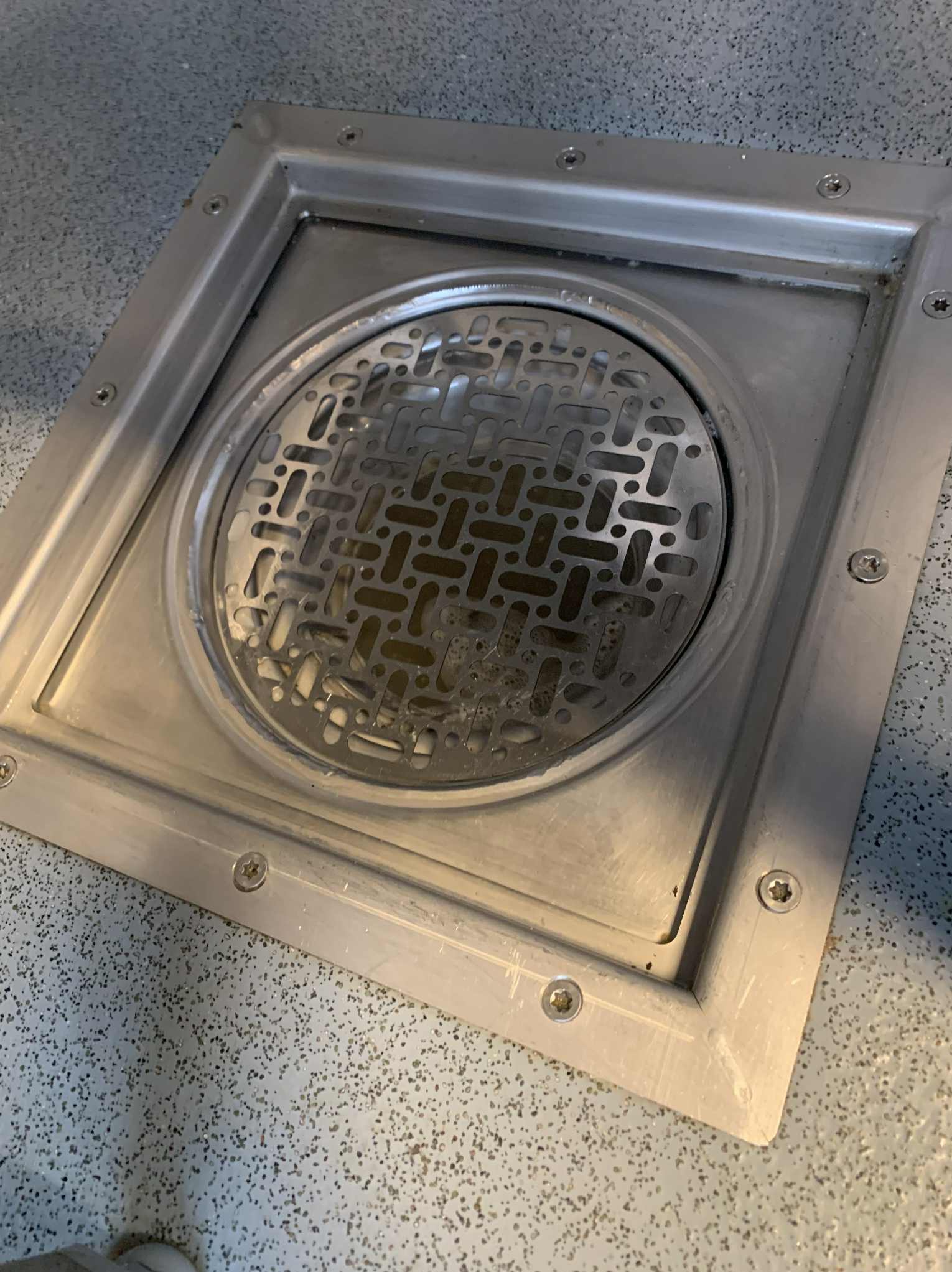 Metalowa kratka ściekowa w podłodze, widoczne zabrudzenia wewnątrz. Kwadratowa rama z widocznymi śrubami mocującymi. Szara, nakrapiana podłoga. Ujęcie z góry.