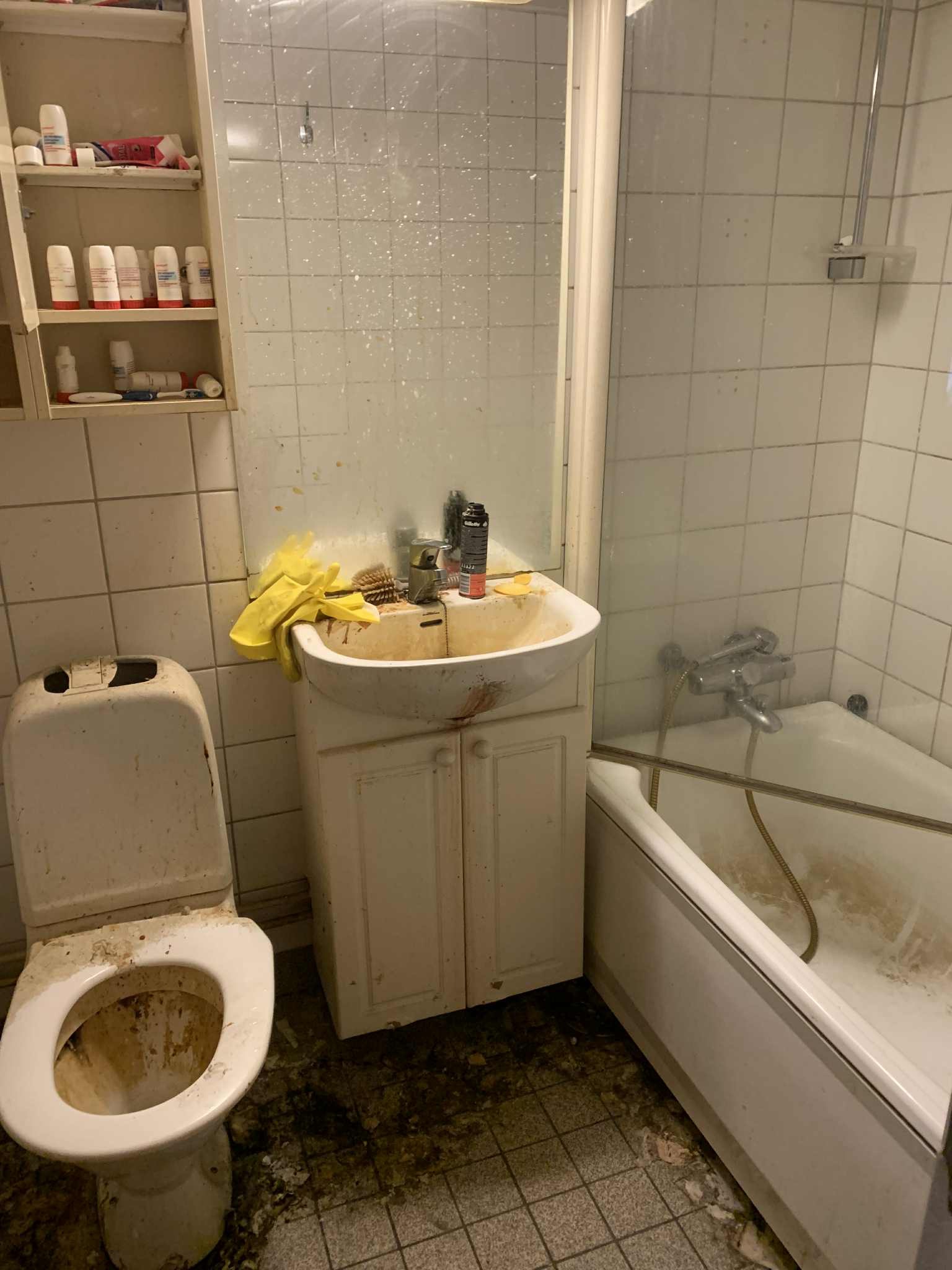 Zaniedbana łazienka z brudną toaletą, umywalką i wanną. Widoczne plamy i zabrudzenia na kafelkach, podłodze i armaturze. Żółte rękawice leżą na umywalce.
