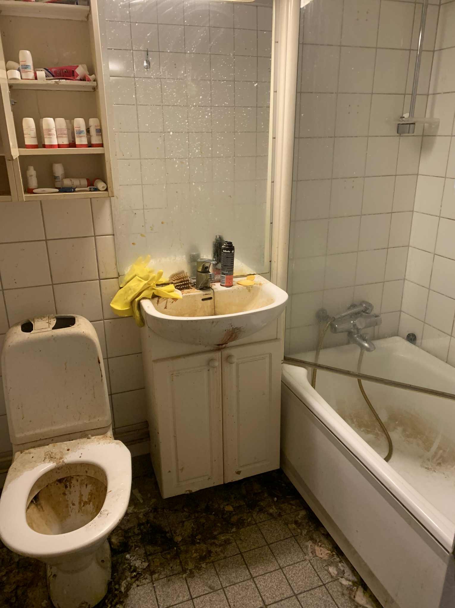Zaniedbana łazienka z brudną toaletą, umywalką i wanną. Żółte rękawice leżą na umywalce. Widoczne plamy i zabrudzenia na podłodze i ścianach.