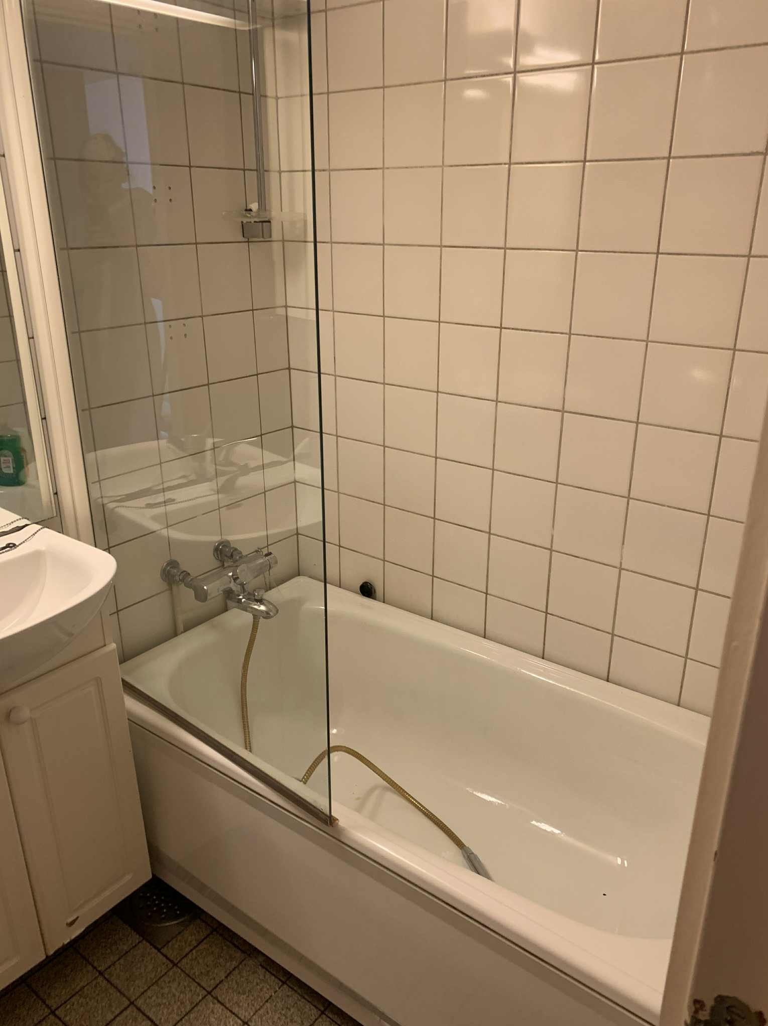 Biała wanna z zamontowanym szklanym parawanem nawannowym, widoczna bateria i słuchawka prysznicowa. Na ścianach jasne, kwadratowe płytki. Umywalka z szafką po lewej.