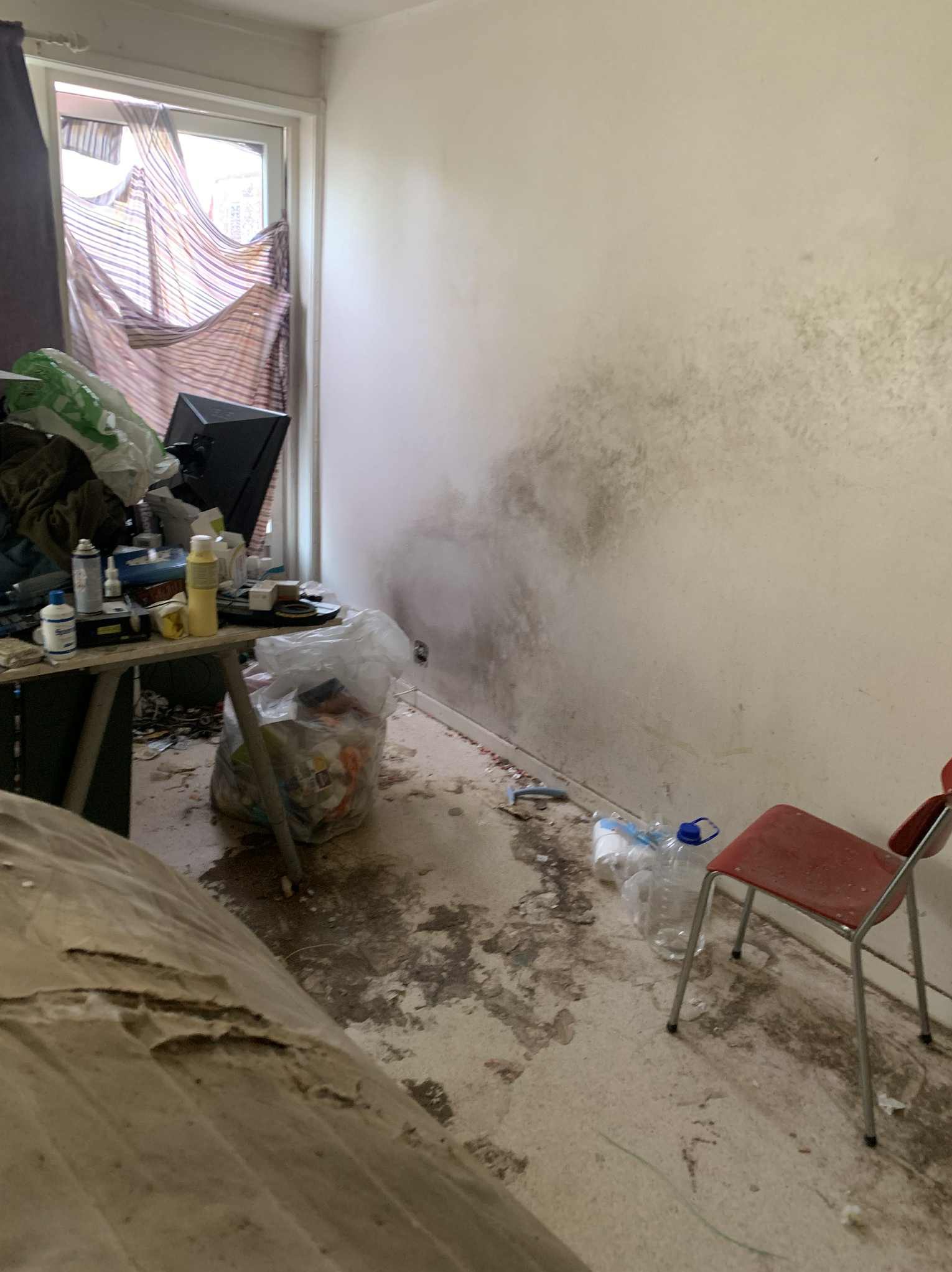 Zaniedbane wnętrze pokoju z widoczną pleśnią na ścianie, zagraconym biurkiem i brudną podłogą. Czerwone krzesło stoi obok plastikowych butelek. Okno zasłonięte materiałem.