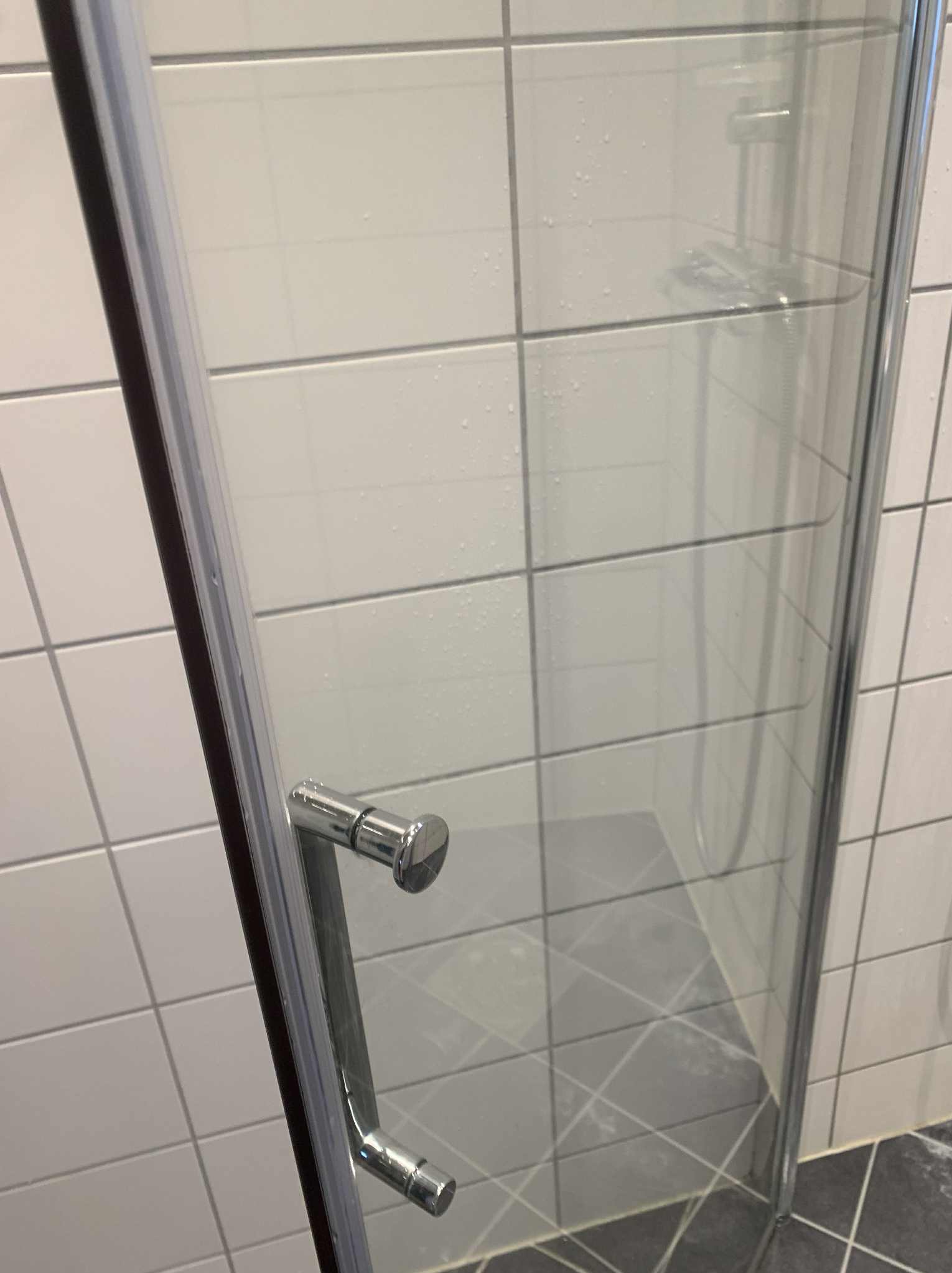 Błyszcząca, chromowana rączka kabiny prysznicowej z widocznymi kroplami wody na szklanej powierzchni i białych kafelkach w tle. Ujęcie z bliska.