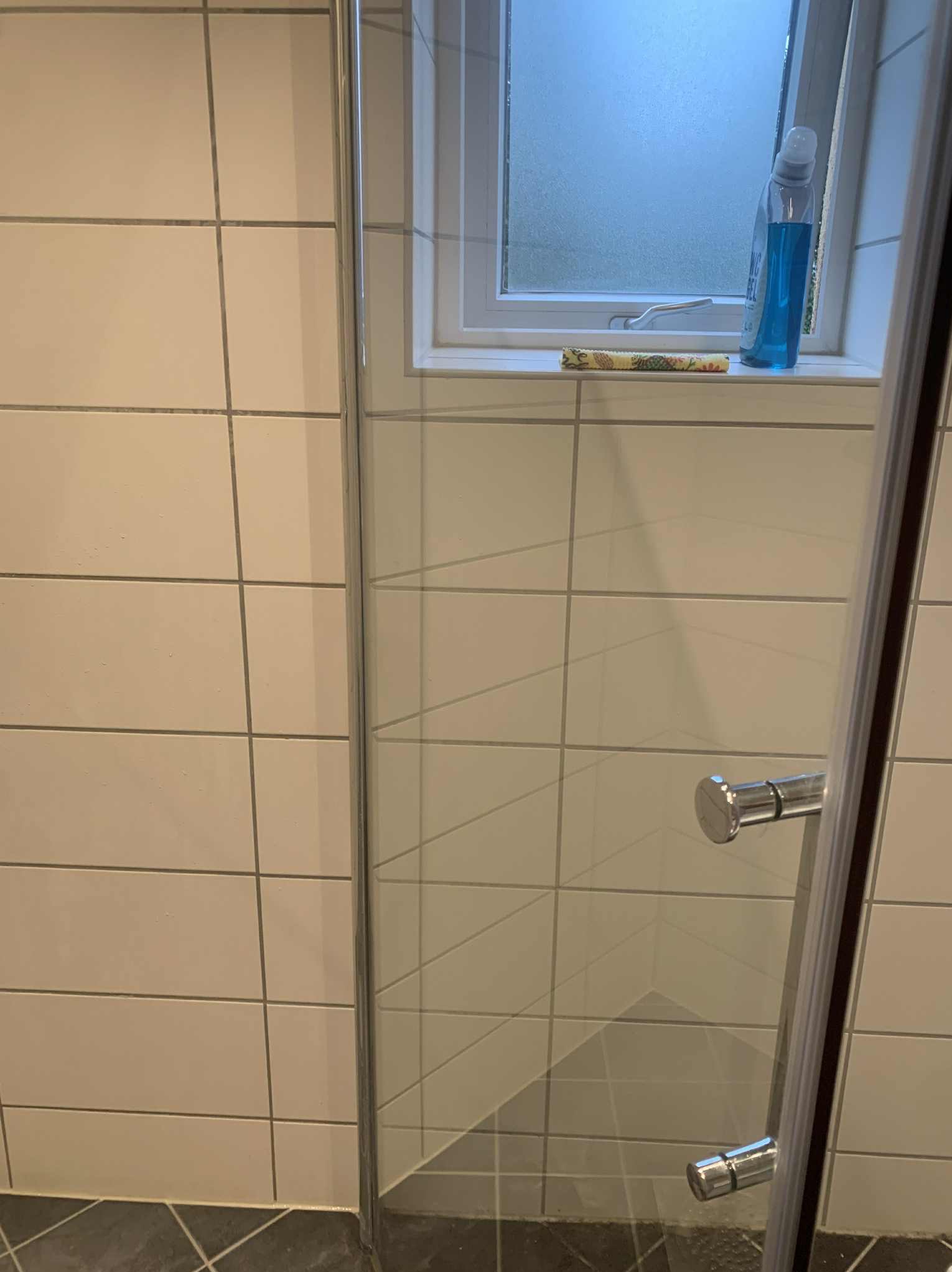 Czysta kabina prysznicowa z widocznymi płytkami i oknem z płynem do czyszczenia, ukazująca efekt sprzątania łazienki.