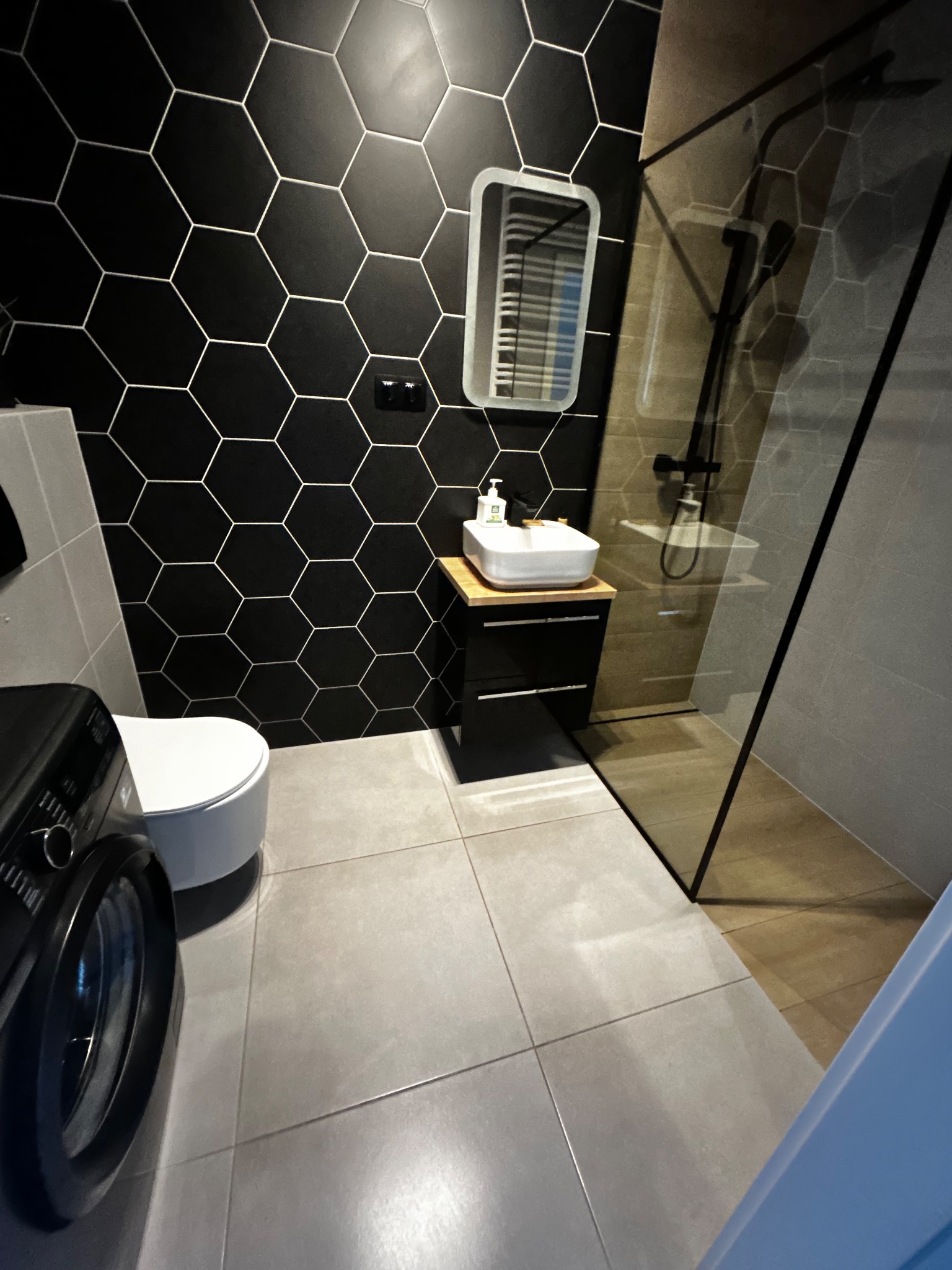 Nowoczesna łazienka z czarnymi heksagonalnymi płytkami, kabiną prysznicową typu walk-in i białą umywalką nablatową. Widoczna pralka i lustro.