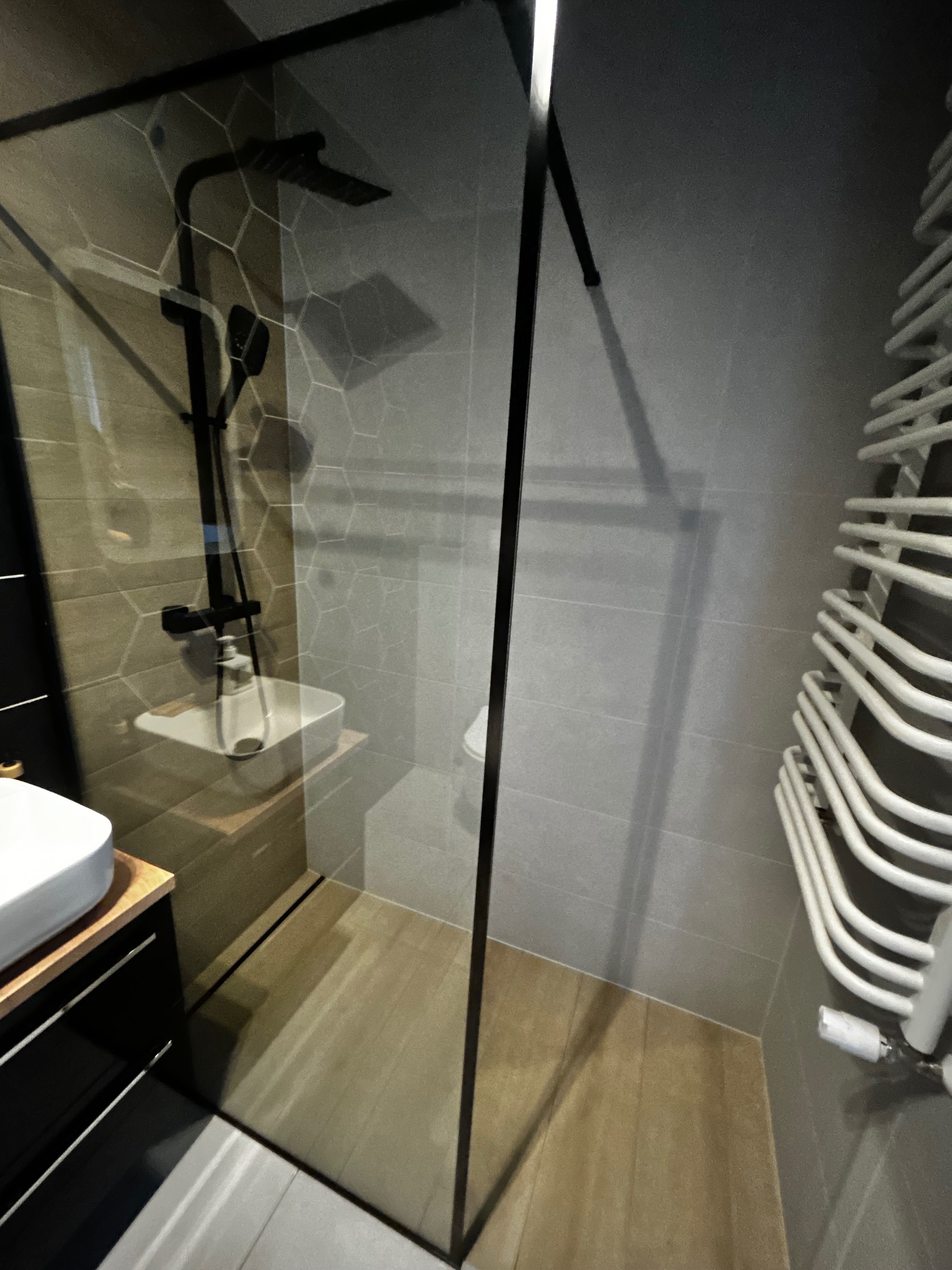 Nowoczesna łazienka z kabiną typu walk-in, czarną armaturą i grzejnikiem drabinkowym. Widoczne płytki heksagonalne i drewnopodobne na podłodze.