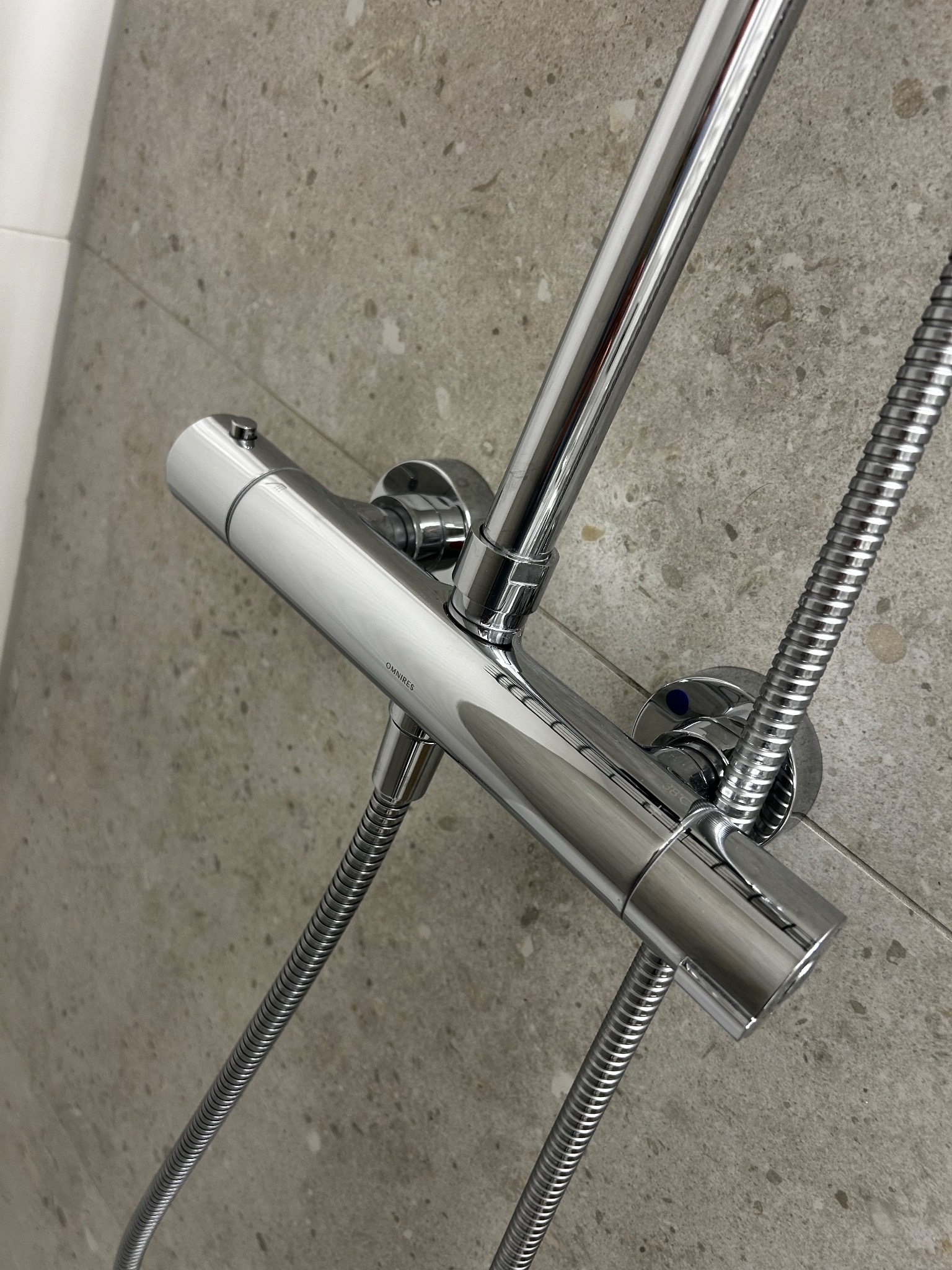 Błyszcząca, chromowana bateria prysznicowa Omnires zamontowana na szarej, kamiennej ścianie. Widoczne węże i głowica prysznicowa. Ujęcie z bliska, z boku.