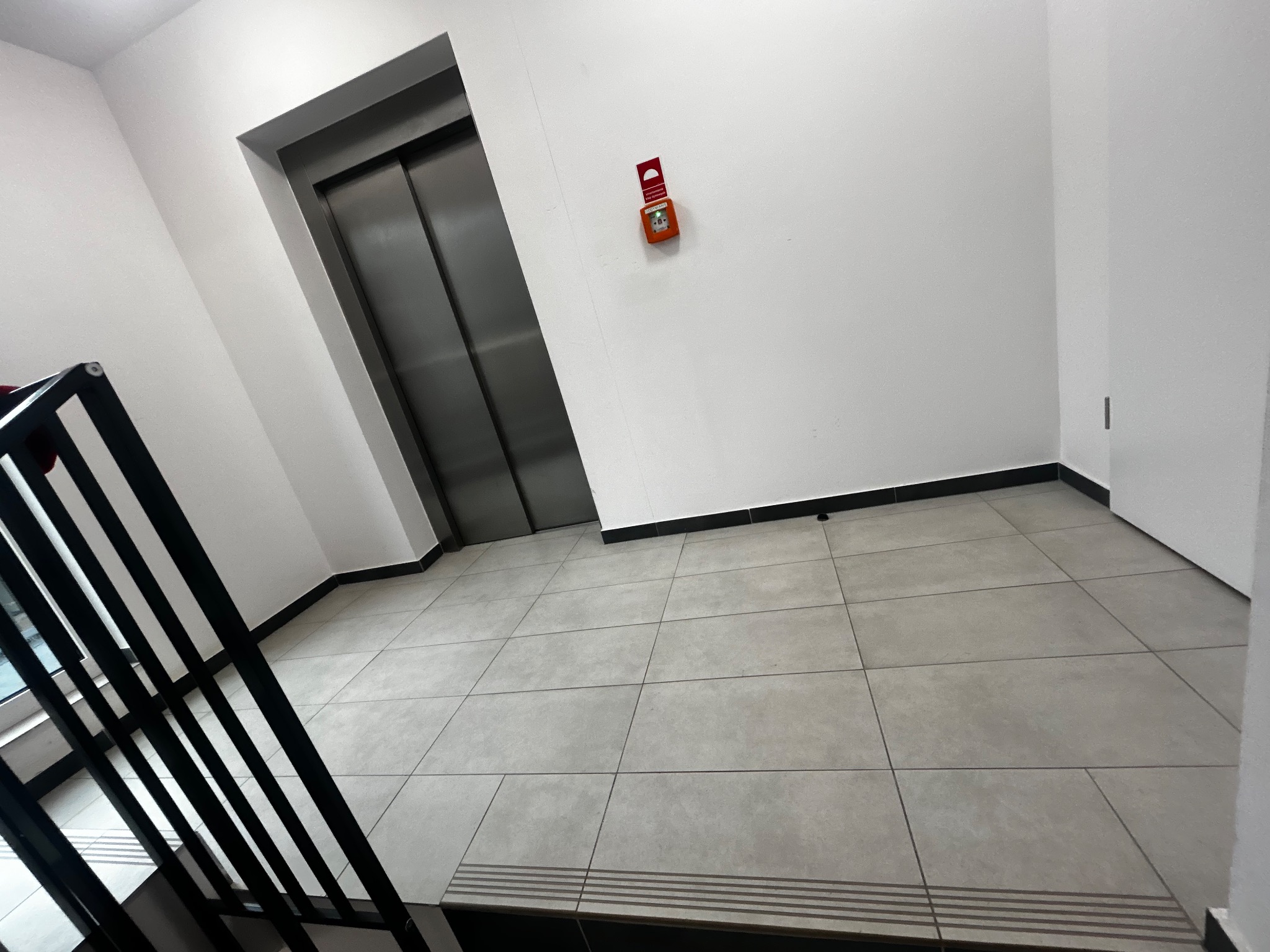 Wnętrze klatki schodowej z szarymi płytkami podłogowymi i metalową windą. Na ścianie widoczny pomarańczowy czujnik dymu. Minimalistyczny design.