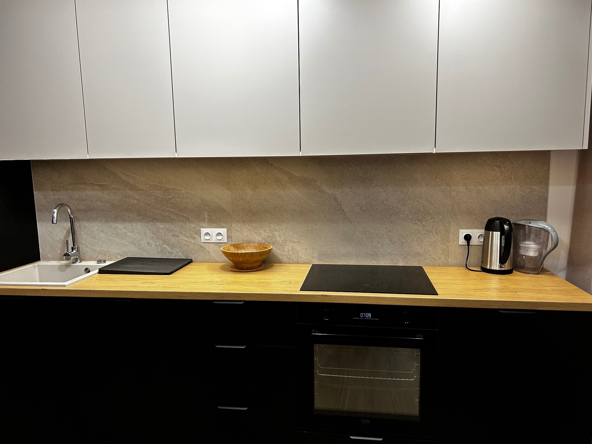 Nowoczesna kuchnia z drewnianym blatem, czarnymi szafkami i białymi szafkami wiszącymi. Na blacie zlewozmywak, płyta indukcyjna, czajnik i misa.