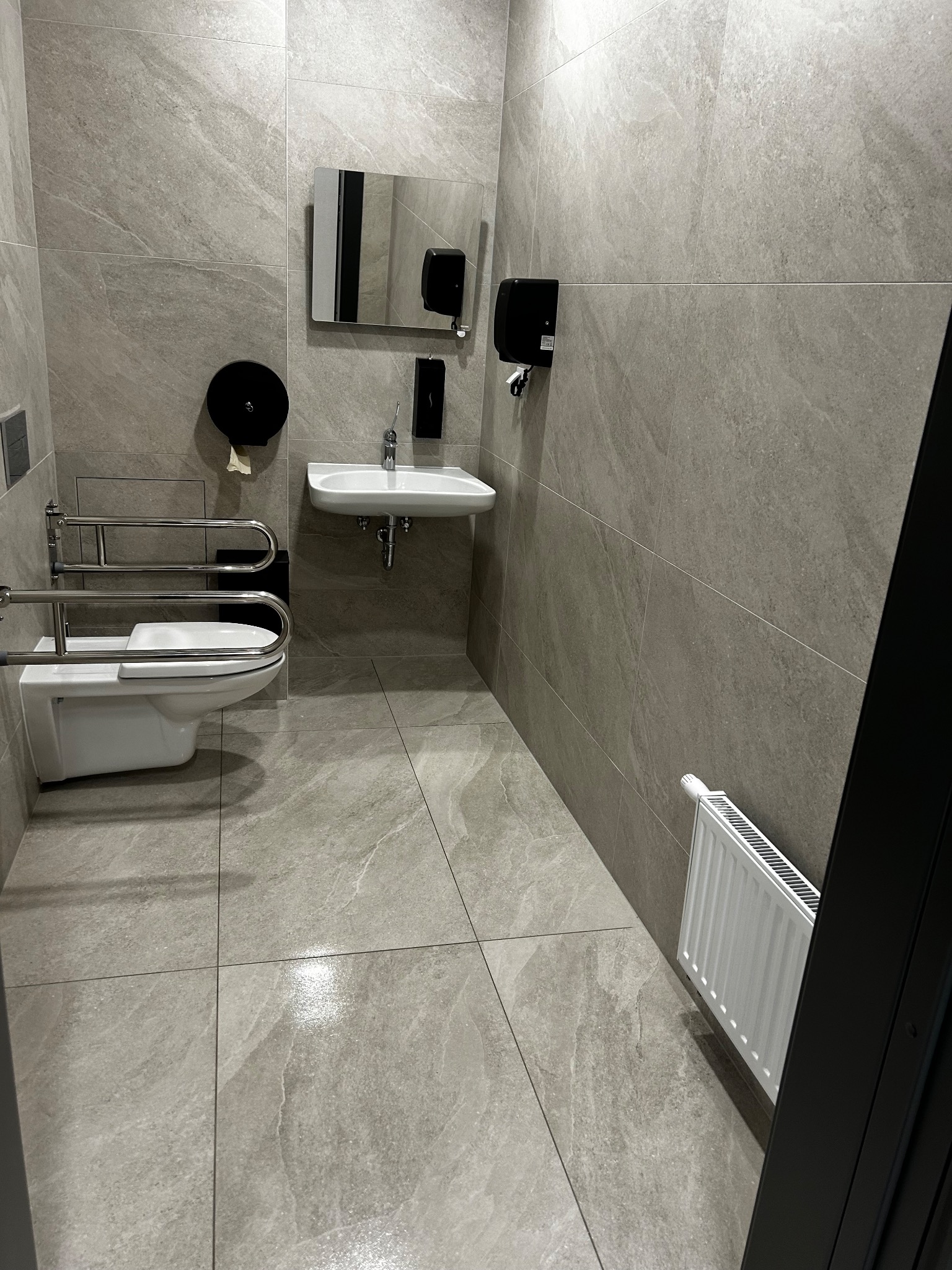 Wnętrze toalety publicznej z udogodnieniami dla niepełnosprawnych: umywalka, toaleta z poręczami, dozownik mydła i papieru, grzejnik. Płytki w odcieniach szarości.