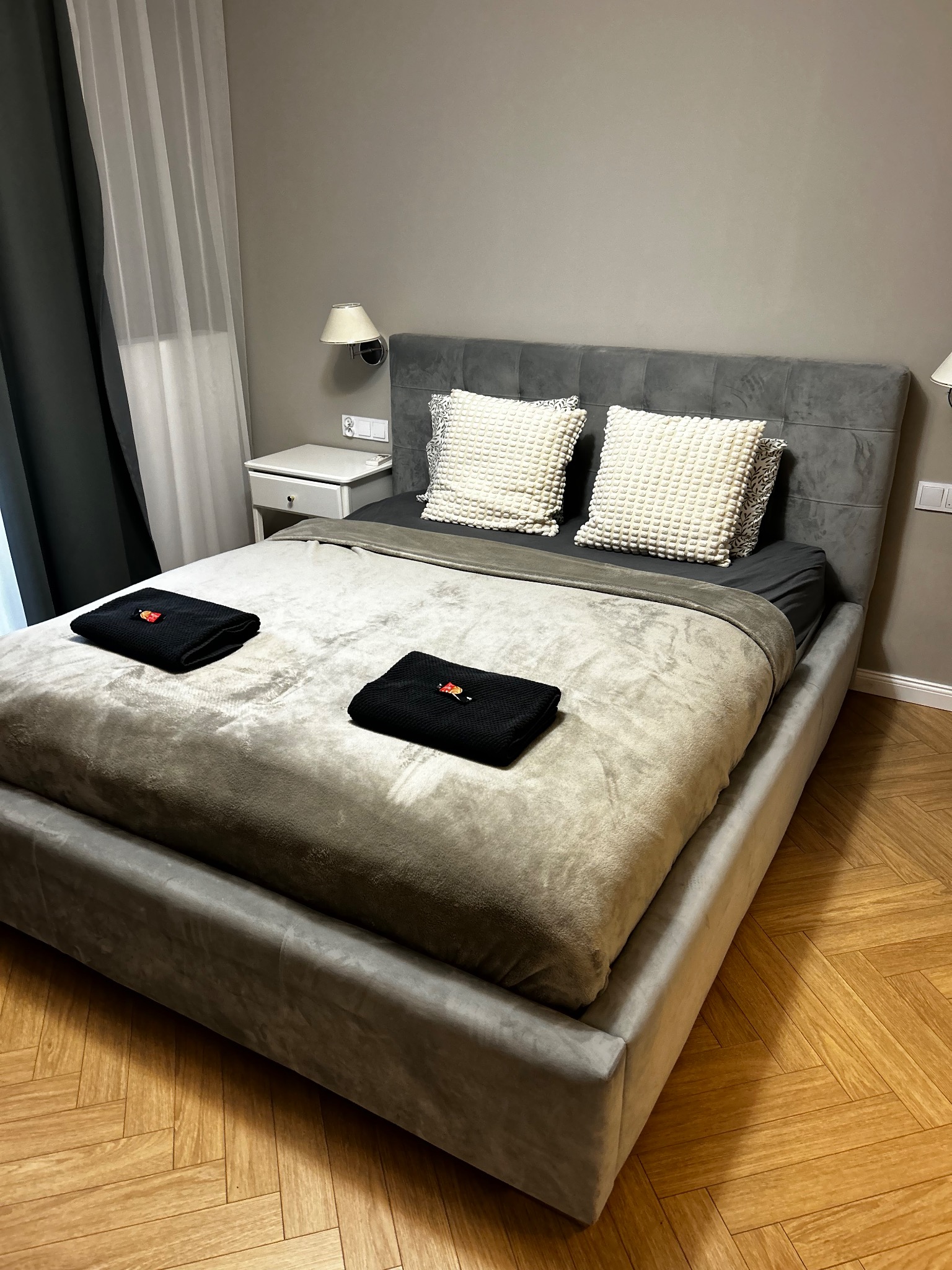 Ustawione łóżko z szarą pościelą, dwoma czarnymi ręcznikami z czerwonym akcentem i dekoracyjnymi poduszkami na tle parkietu i ściany w odcieniach szarości.