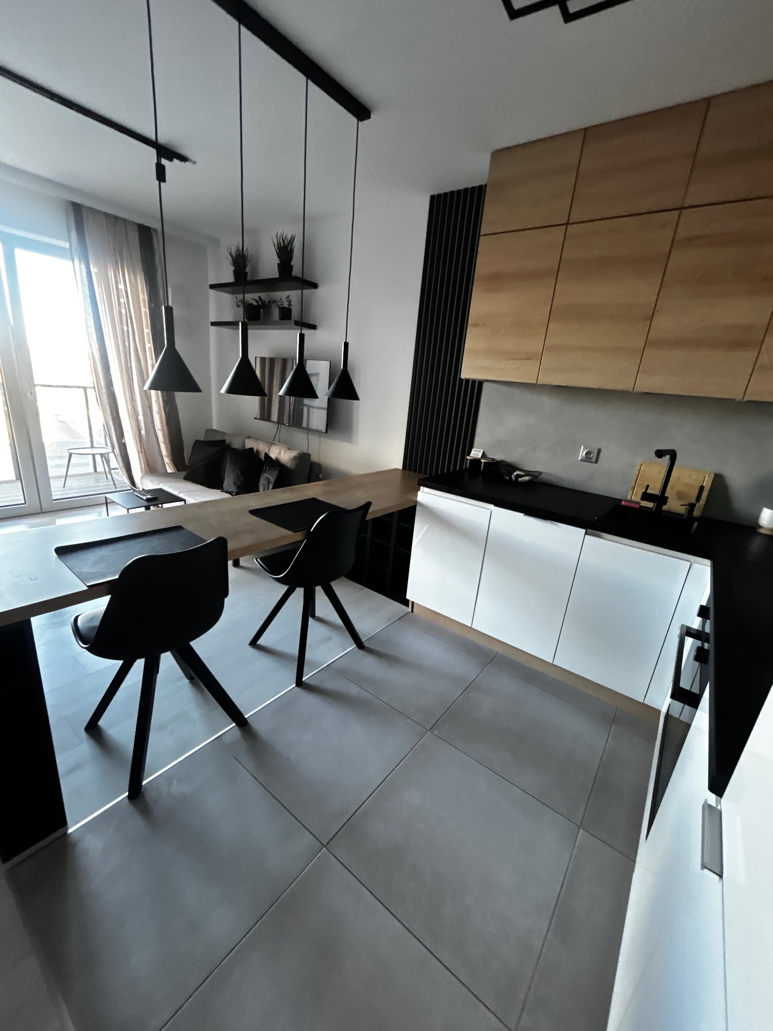 Nowoczesne wnętrze: kuchnia z drewnianymi szafkami, czarnym blatem i białymi frontami, połączona z salonem. Widoczne czarne krzesła i lampy.
