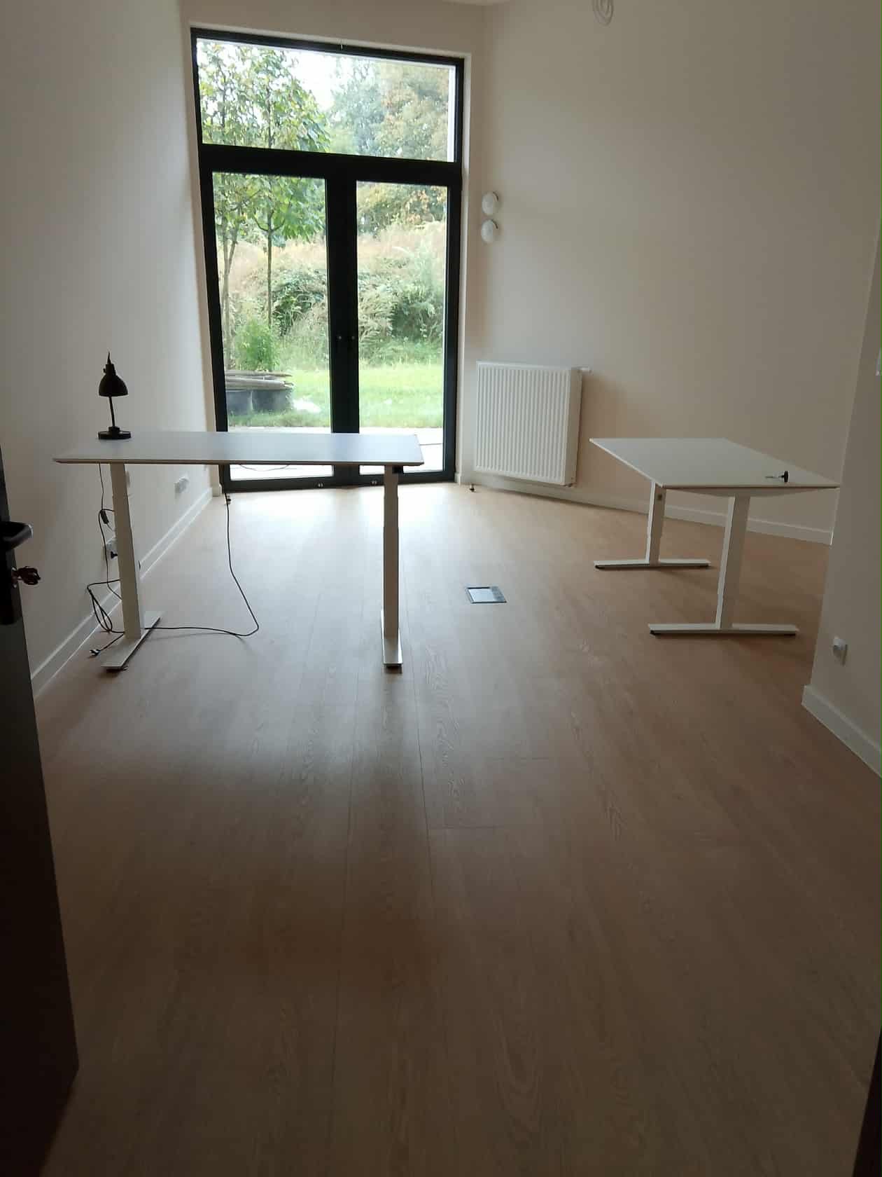 Puste biuro we Wrocławiu po sprzątaniu. Dwa białe biurka, duże okno z widokiem na zieleń, jasne ściany i podłoga. Minimalistyczny design, czysta przestrzeń.