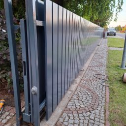 Air-Send - Nowoczesna, przesuwna brama wjazdowa z metalowymi panelami w odcieniach szarości, zamontowana na posesji z kostką brukową i zielenią w tle.