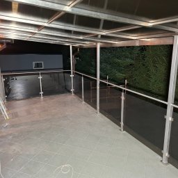 Air-Send - Zadaszony taras z balustradą szklaną i metalowymi elementami konstrukcyjnymi. Widoczna drabina, sugerująca niedawny montaż. Tło stanowią ciemne drzewa.