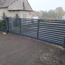 Air-Send - Nowoczesna, pozioma brama wjazdowa z metalu w kolorze grafitowym, z widocznym mechanizmem przesuwnym, na tle wiejskiego krajobrazu z budynkiem gospodarczym i drzewami.