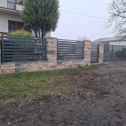 Air-Send - Nowoczesna brama wjazdowa z poziomych, szarych profili, osadzona na słupkach z dekoracyjnej, warstwowej kostki. Tło: budynek i drzewo bez liści.