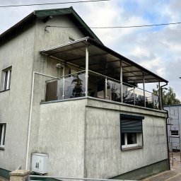Air-Send - Zadaszony balkon z przeszkloną balustradą na piętrze domu. Widoczne elementy konstrukcyjne i zadaszenie z poliwęglanu. Ujęcie z dołu, pochmurne niebo w tle.