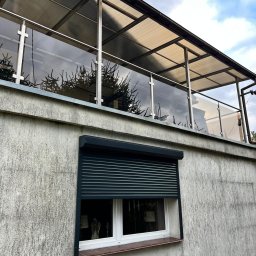 Air-Send - Szklana balustrada balkonowa z elementami ze stali nierdzewnej, widoczna z dołu, z roletą okienną w tle. Nowoczesny design zadaszenia.