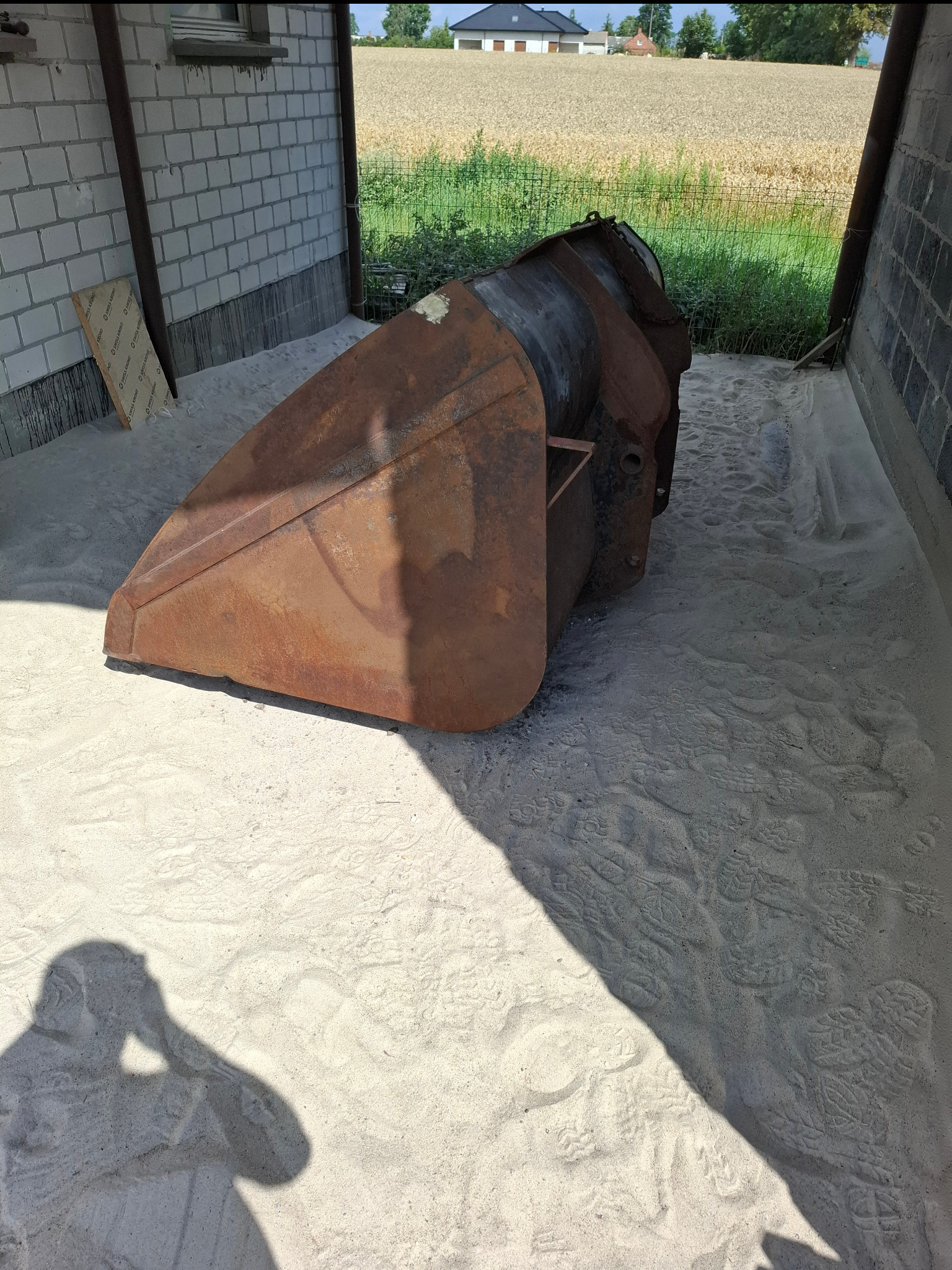 Zardzewiała łyżka koparki w piasku, widoczny cień osoby. W tle budynek i pole. Sprzęt budowlany w trakcie renowacji lub przygotowania do piaskowania.