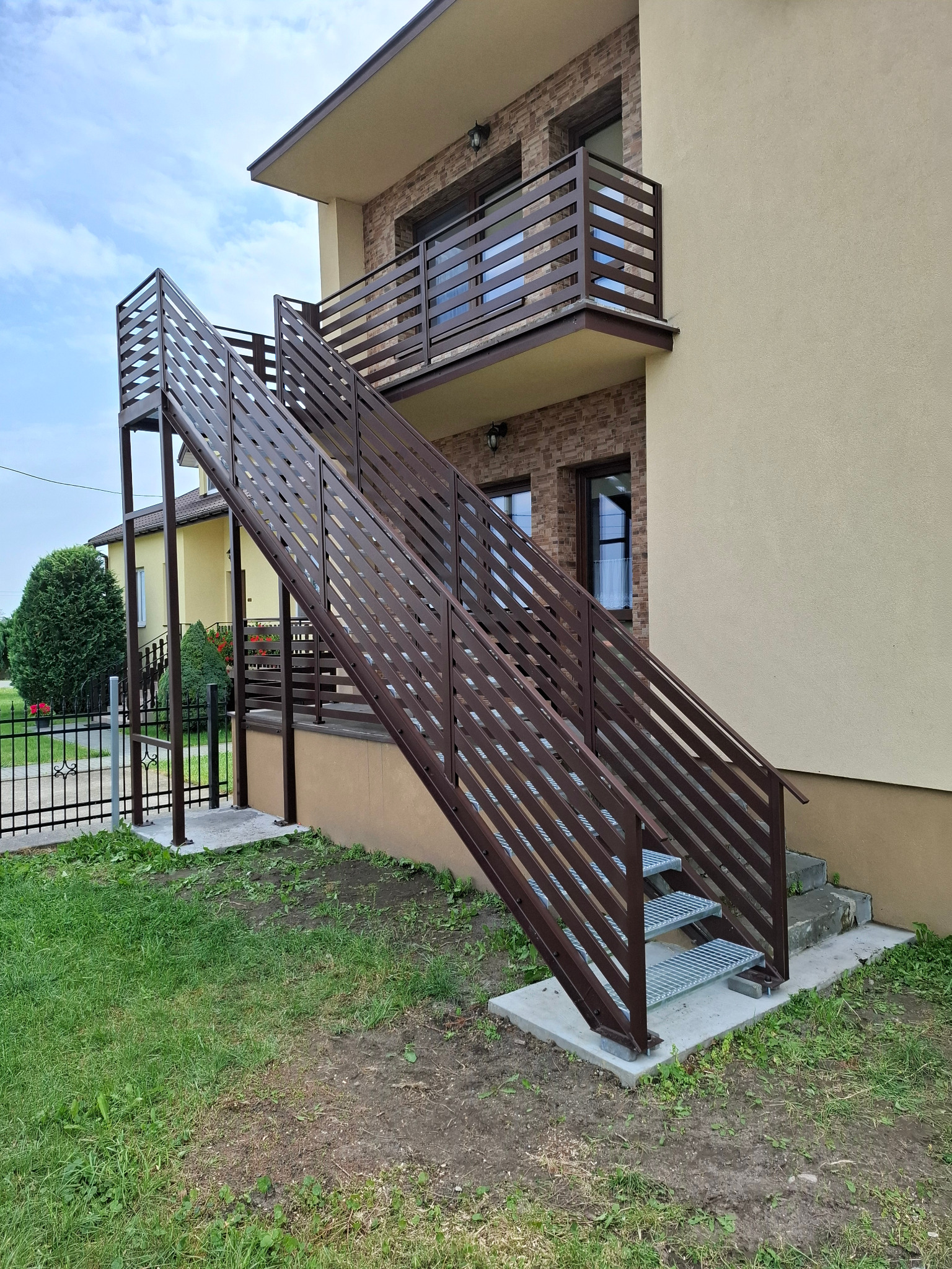 Zewnętrzne schody metalowe z brązową balustradą prowadzące na balkon. Widoczne elementy konstrukcyjne i ażurowe stopnie. Tło stanowi elewacja budynku.