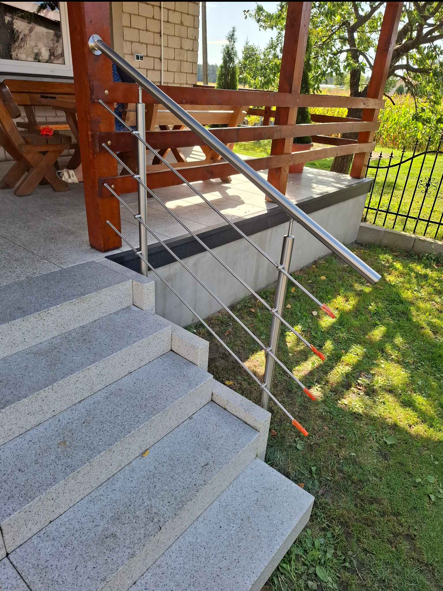 Zewnętrzne schody z jasnego kamienia z nowoczesną, metalową balustradą z cienkich prętów. Widok z góry na schody i fragment ogrodu z zieloną trawą.