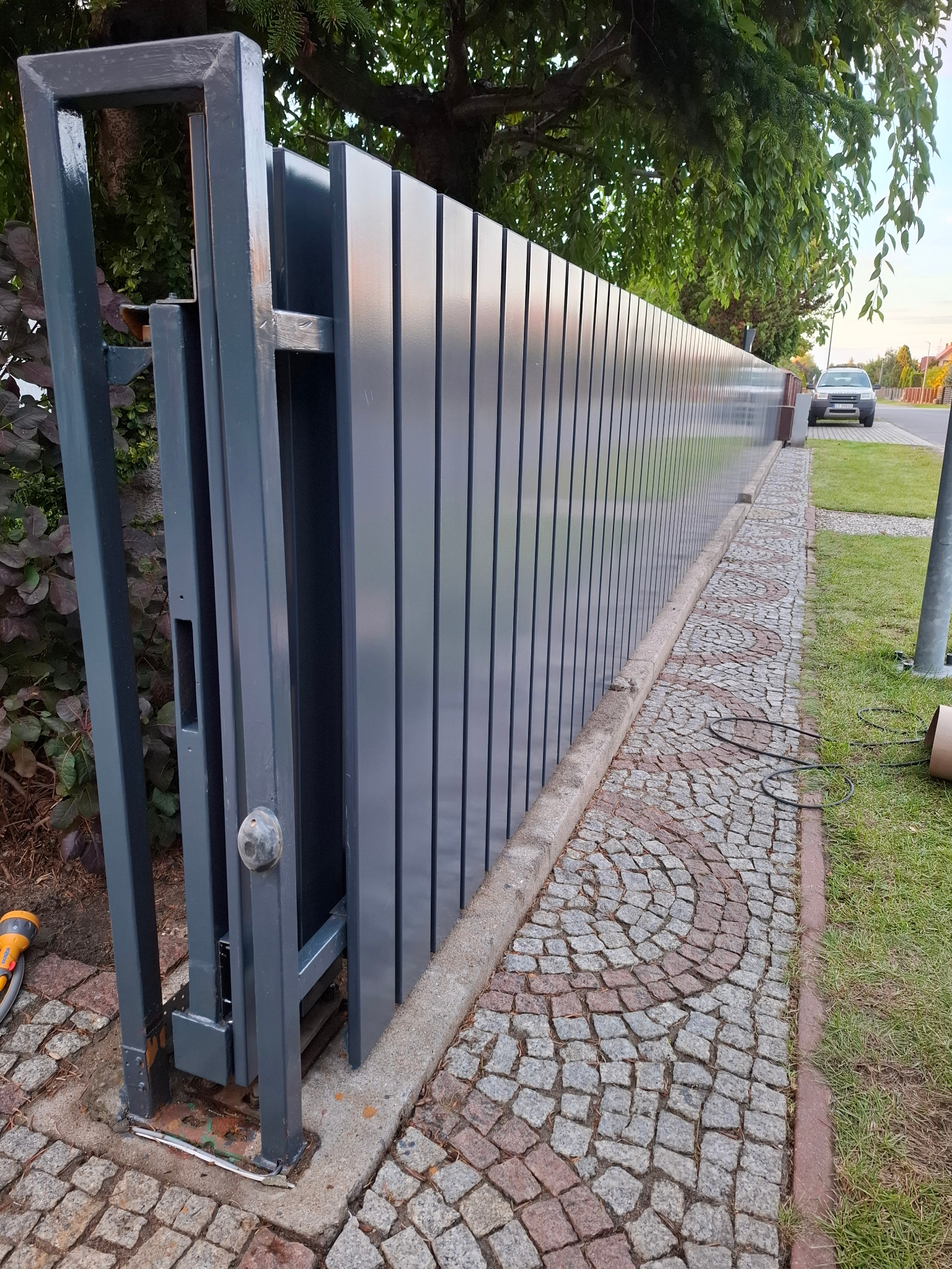 Nowoczesna, przesuwna brama wjazdowa z metalowymi panelami w odcieniach szarości, zamontowana na posesji z kostką brukową i zielenią w tle.