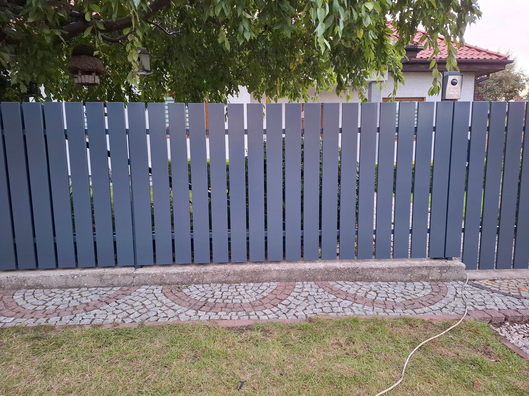 Nowoczesne, szare ogrodzenie panelowe z pionowych desek, widoczne na tle zieleni trawnika i drzew. Uwagę przykuwa dekoracyjny chodnik z kostki brukowej u podstawy.