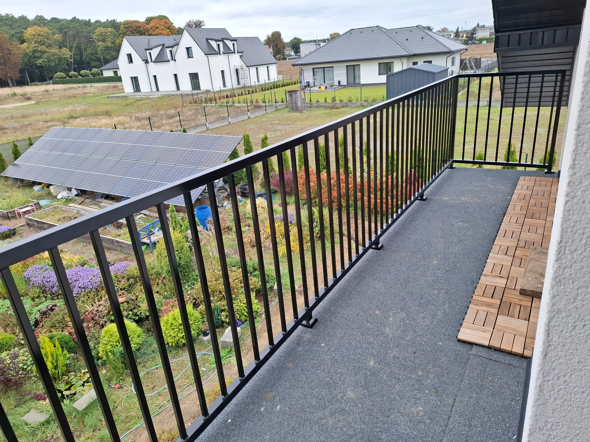 Czarna, metalowa balustrada balkonowa z widokiem na ogród i domy w tle. Elementy drewniane na podłodze balkonu. Ujęcie z góry, podkreślające detale konstrukcji.