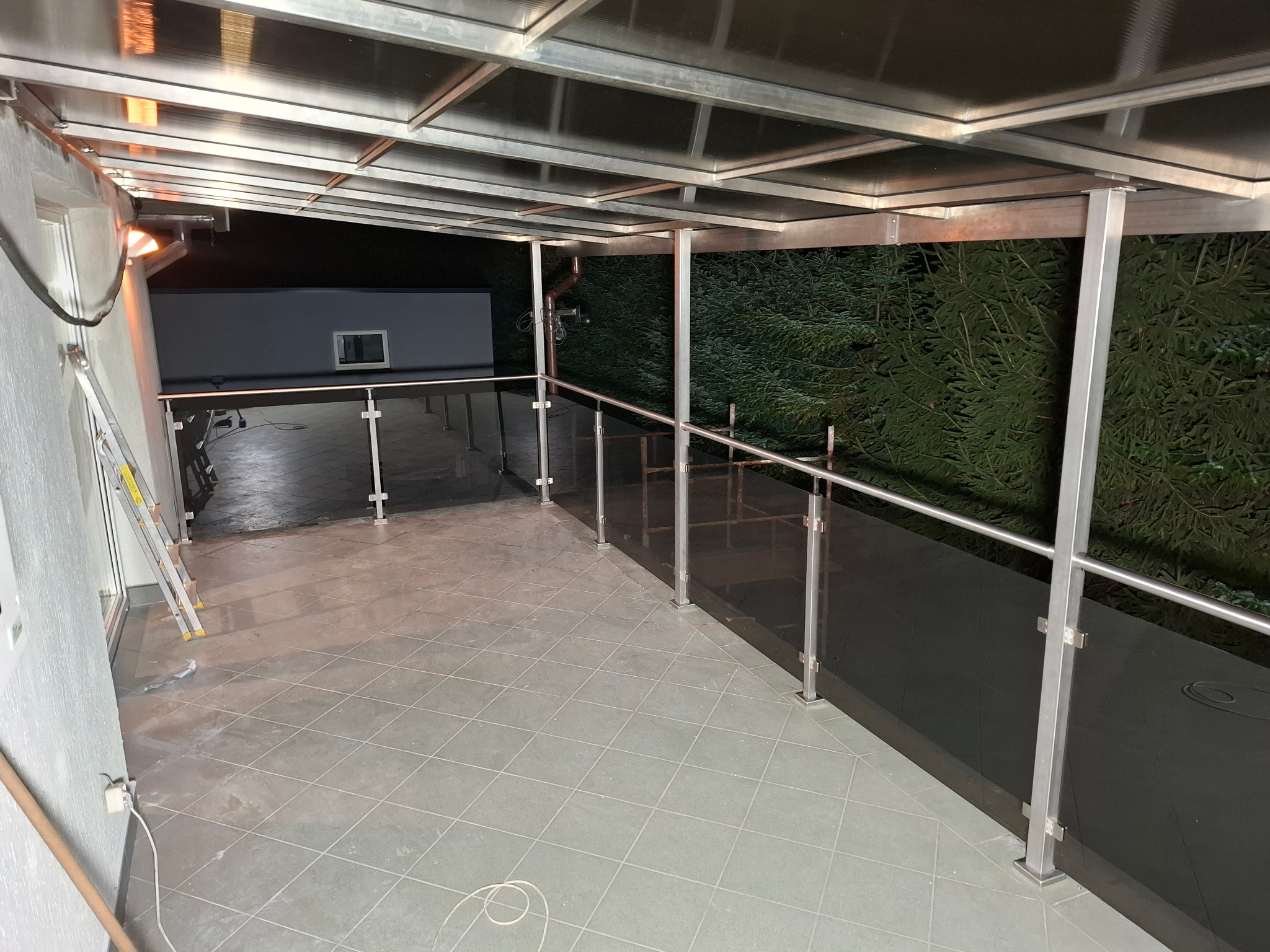 Zadaszony taras z balustradą szklaną i metalowymi elementami konstrukcyjnymi. Widoczna drabina, sugerująca niedawny montaż. Tło stanowią ciemne drzewa.