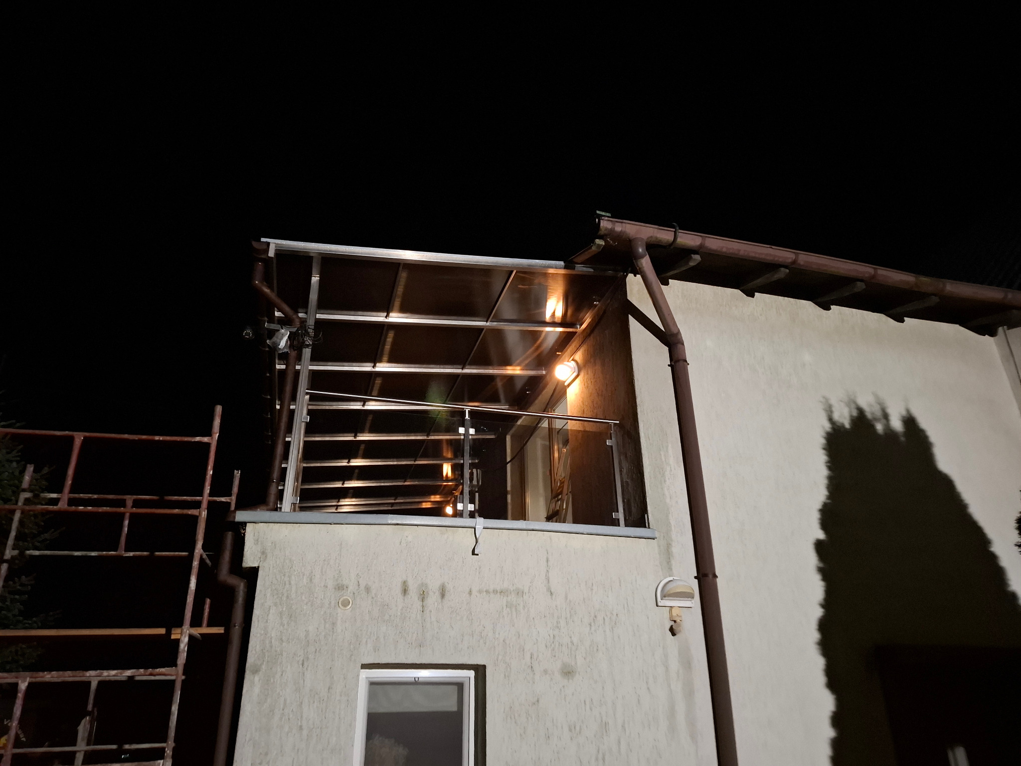 Oświetlony balkon z przeszkloną balustradą i zadaszeniem z poliwęglanu, widoczny z dołu nocą, z fragmentem rusztowania po lewej stronie.