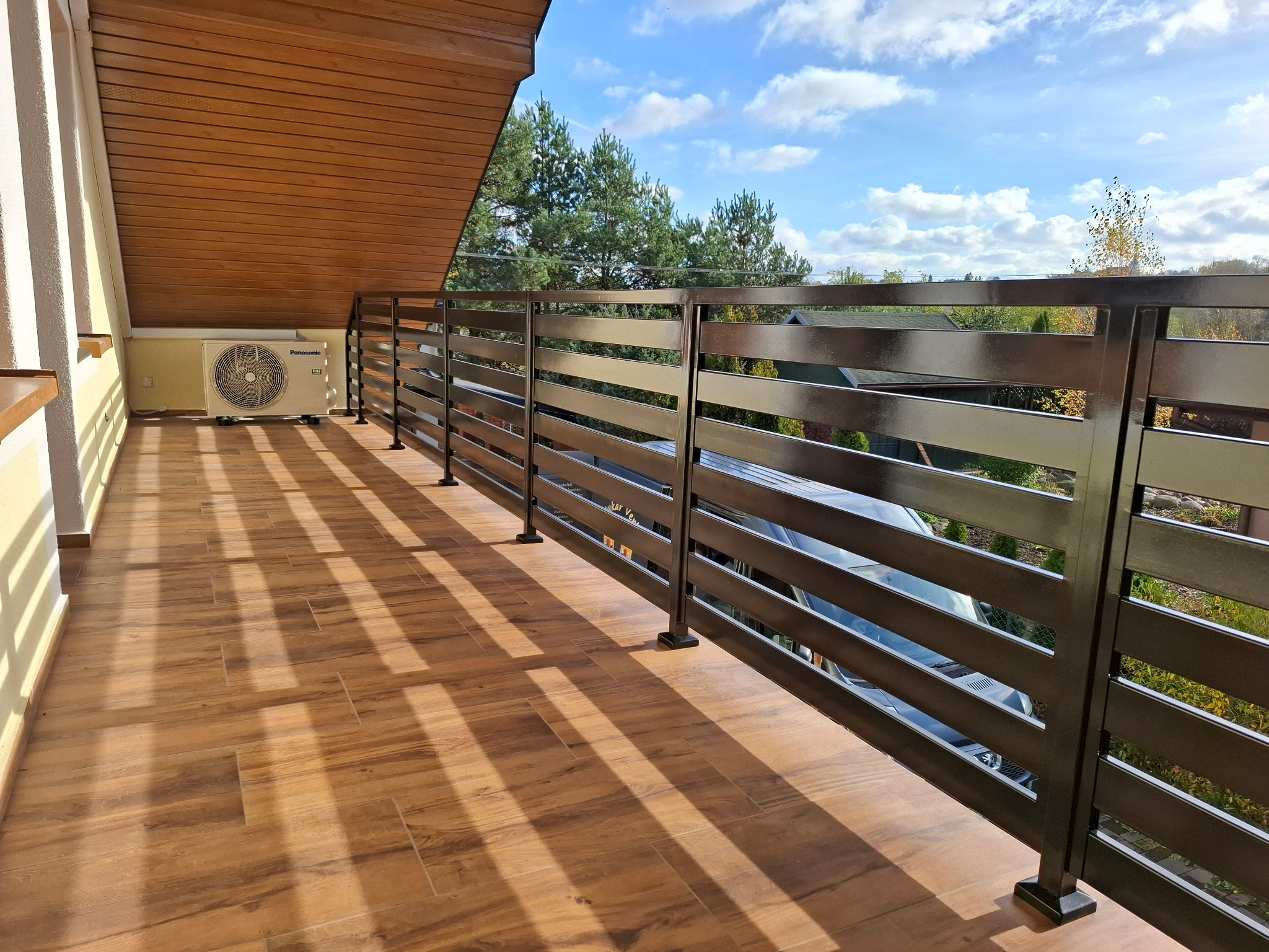 Balkon z poziomą, metalową balustradą w kolorze grafitowym, z widokiem na zieleń i błękitne niebo. Podłoga wyłożona płytkami imitującymi drewno.