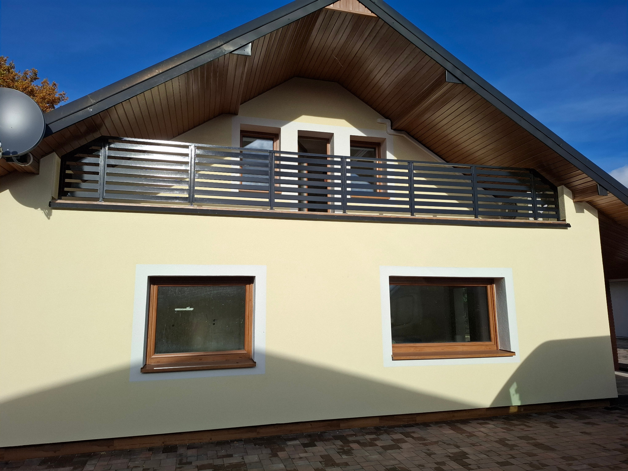 Elewacja domu z balkonem zabezpieczonym nowoczesną, poziomą, metalową konstrukcją. Widoczne dwa okna na parterze i drewniane wykończenie dachu.