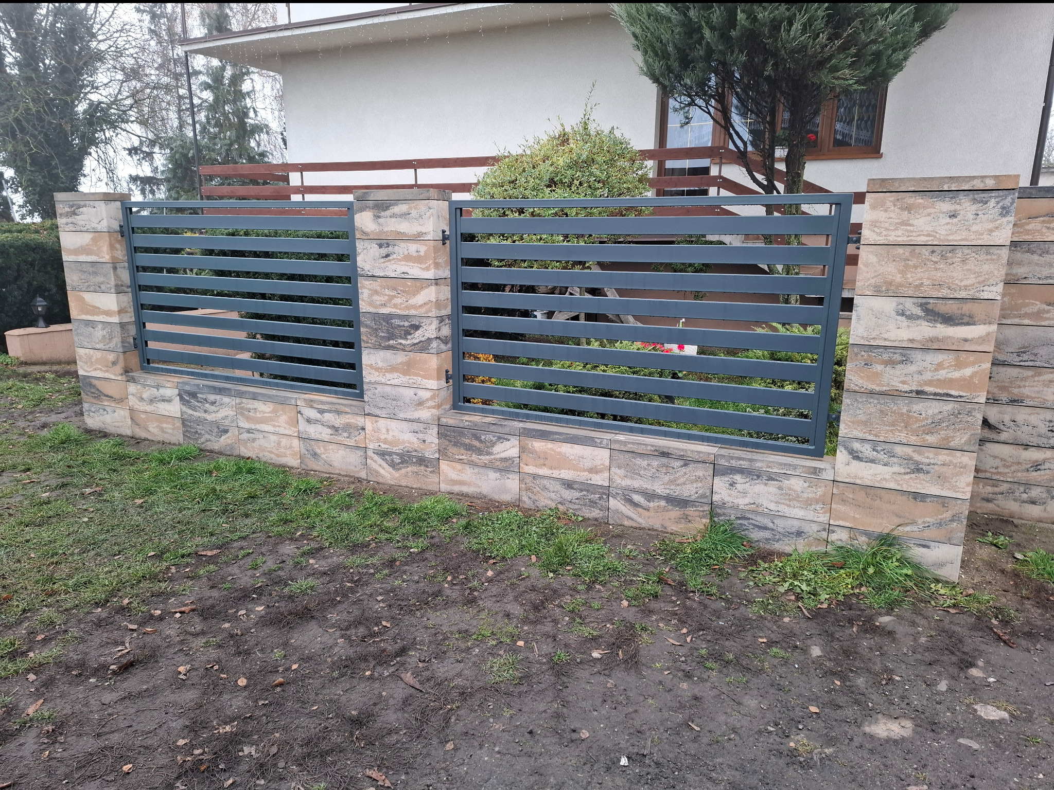 Nowoczesne ogrodzenie panelowe z szarego metalu, osadzone na słupkach z dekoracyjnej kostki. Widok na dom i zieleń w tle. Ujęcie z poziomu gruntu.