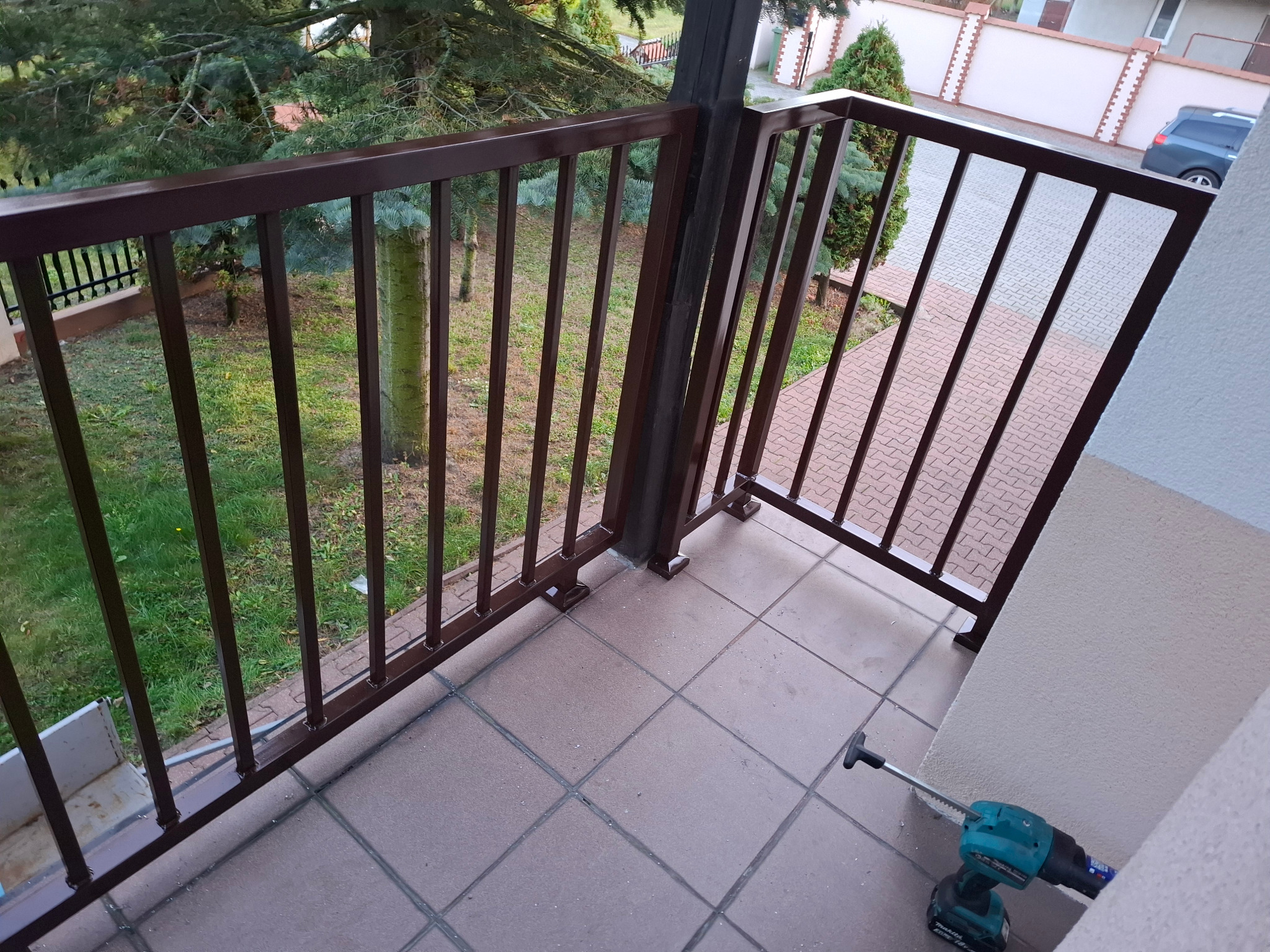 Brązowa, metalowa balustrada balkonowa z widokiem na zieleń i narzędziem (wiertarką) w rogu. Ujęcie z góry, podkreślające detale konstrukcji i otoczenie.