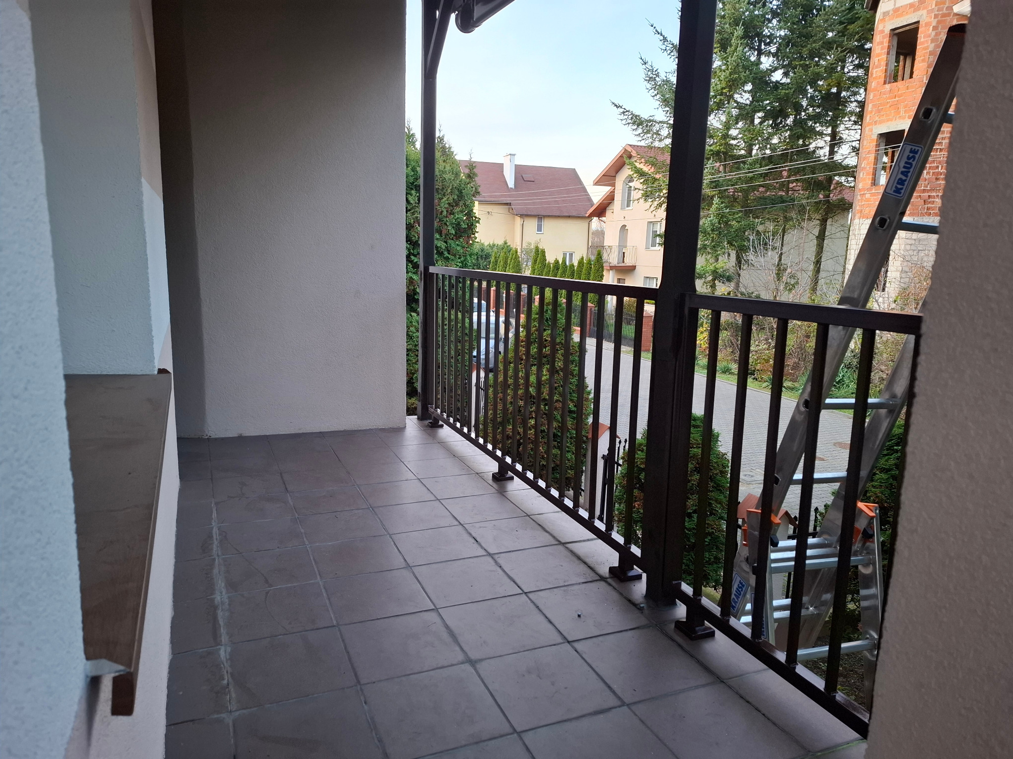 Balkon z brązową, metalową balustradą na tle zieleni i budynków. Na balkonie widoczna drabina Krause. Ujęcie z wnętrza balkonu, z widokiem na zewnątrz.